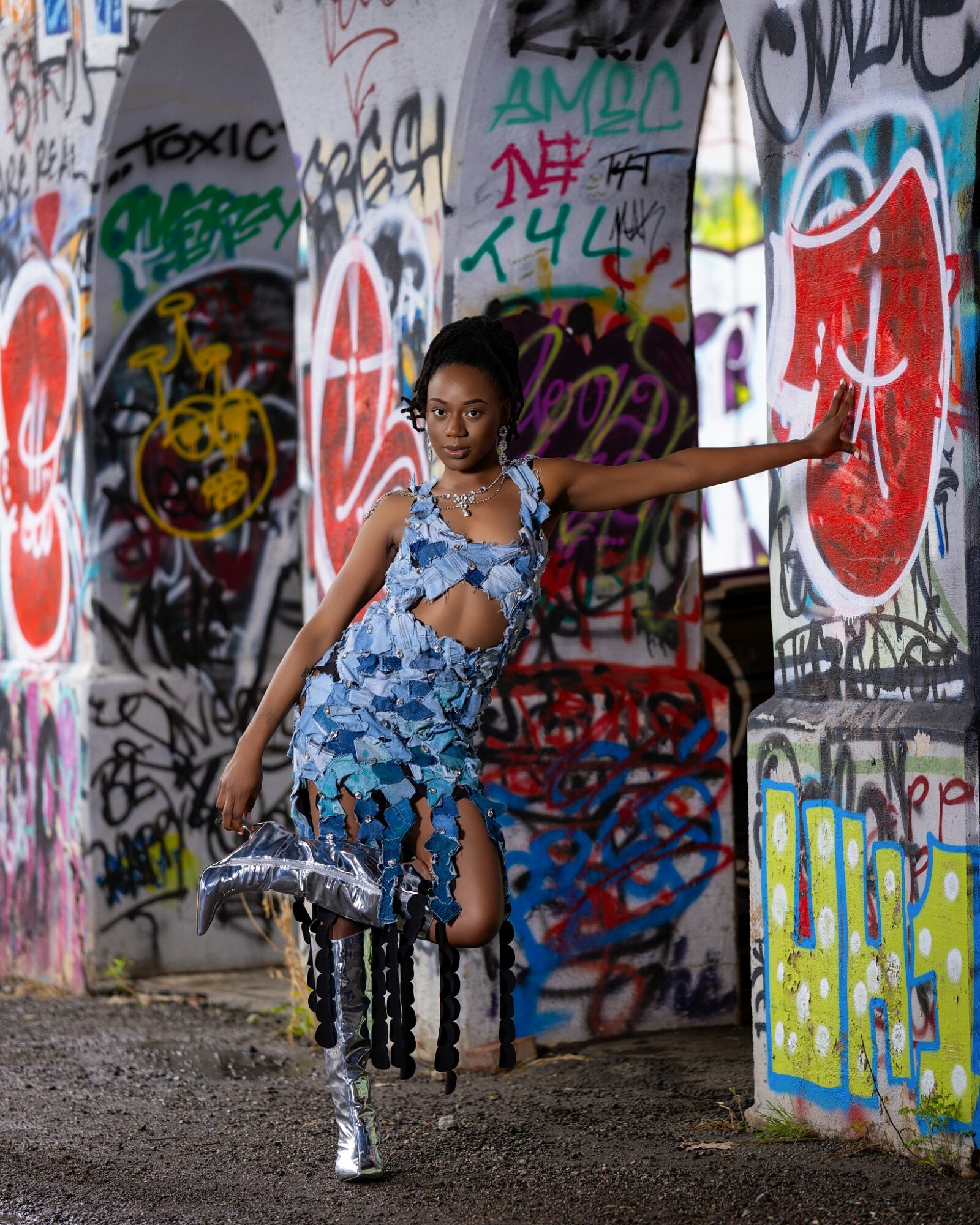 Young woman in a blue patterned outfit poses with graffiti-covered walls, holding a bag, extending her arm to the side.