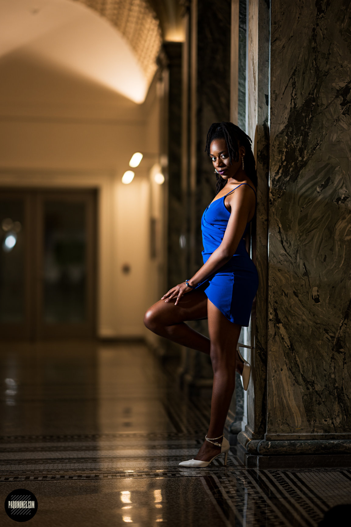Woman in a blue dress leaning against a marble wall in an elegant interior, dimly lit with warm lighting.