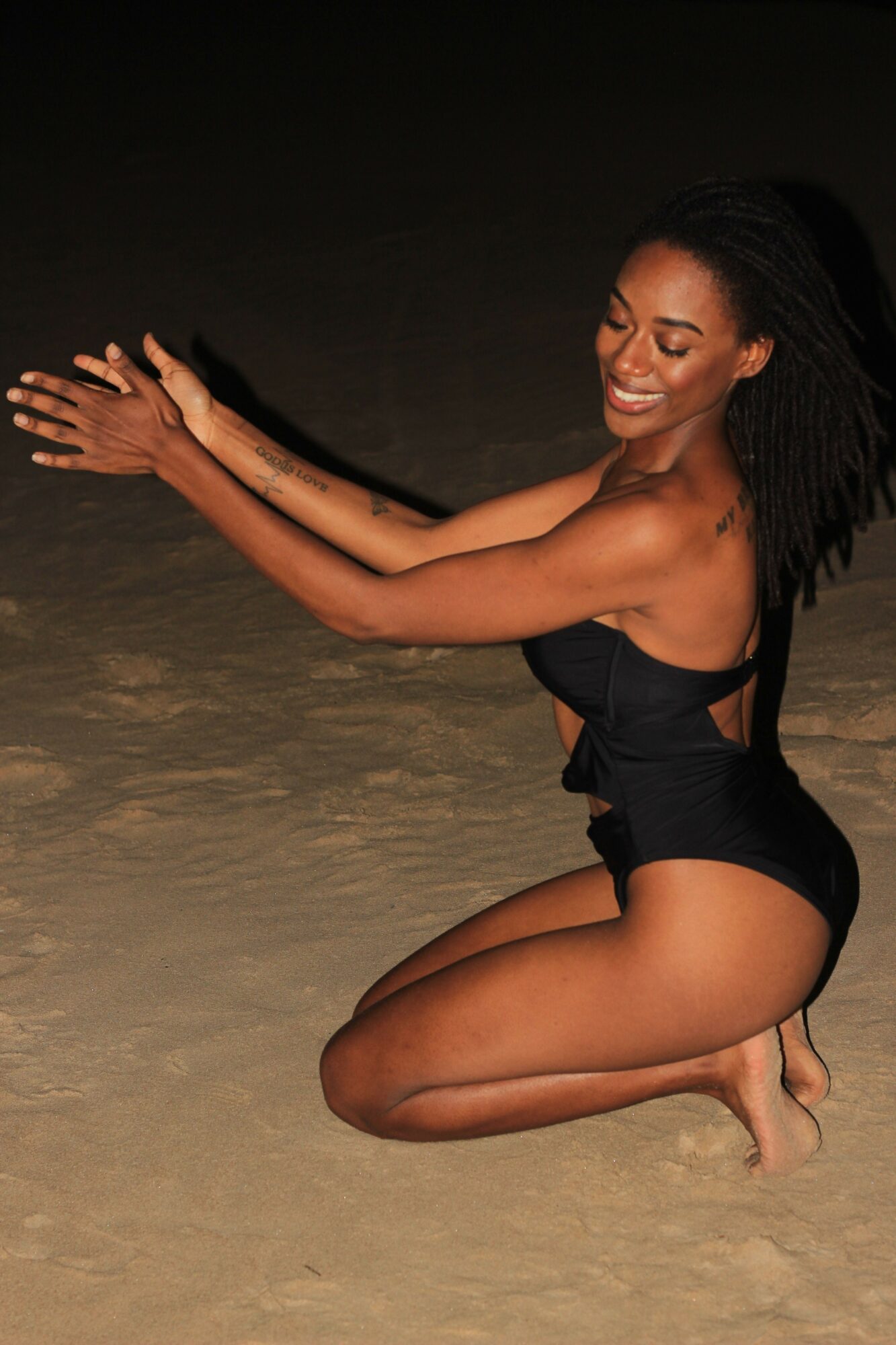 Woman lying on her side with arms raised, smiling, wearing black clothing, dark background, sandy surface.