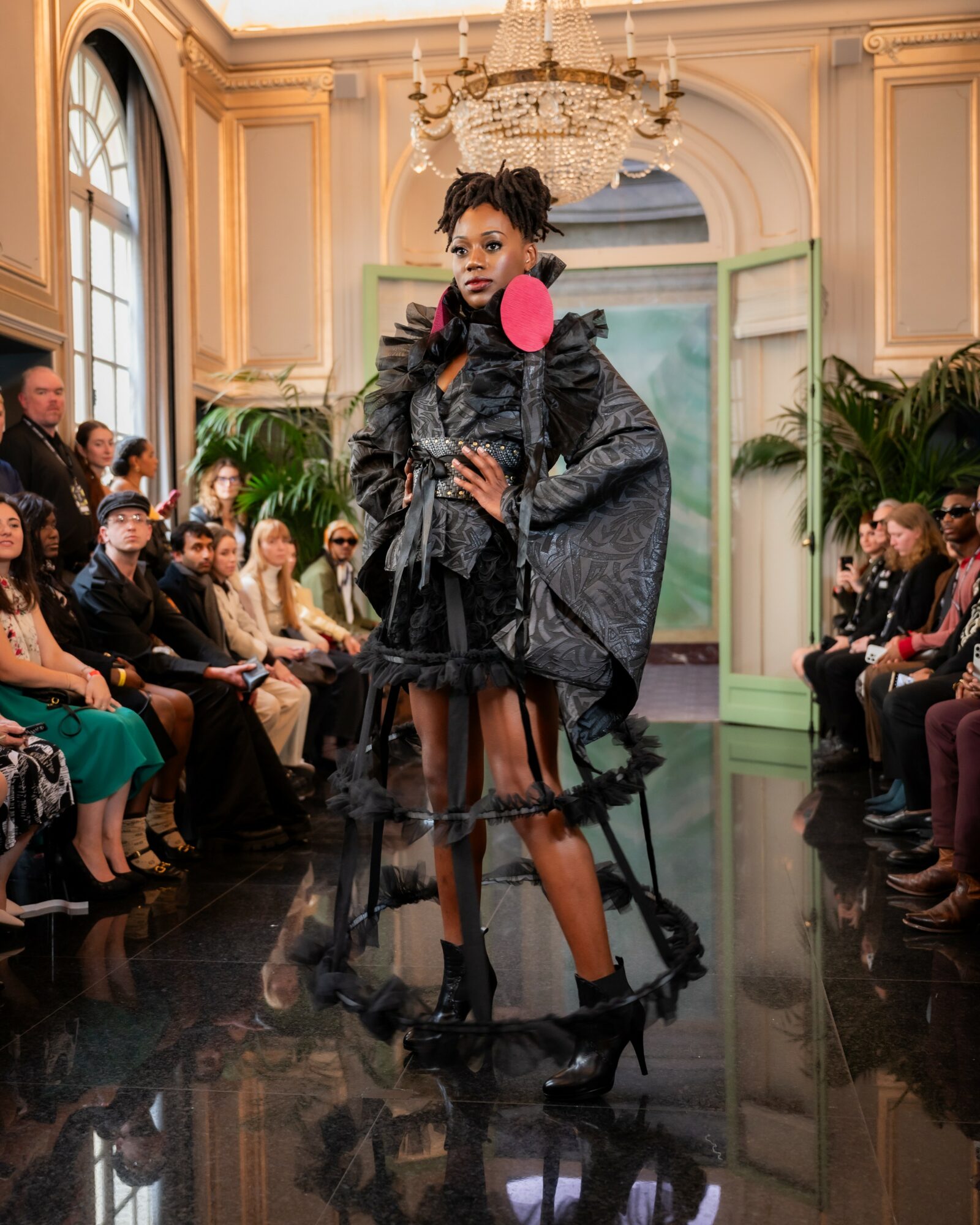 Model walking runway in a grand room with chandeliers and audience, wearing a dark, layered outfit with pink accessory, high heels.