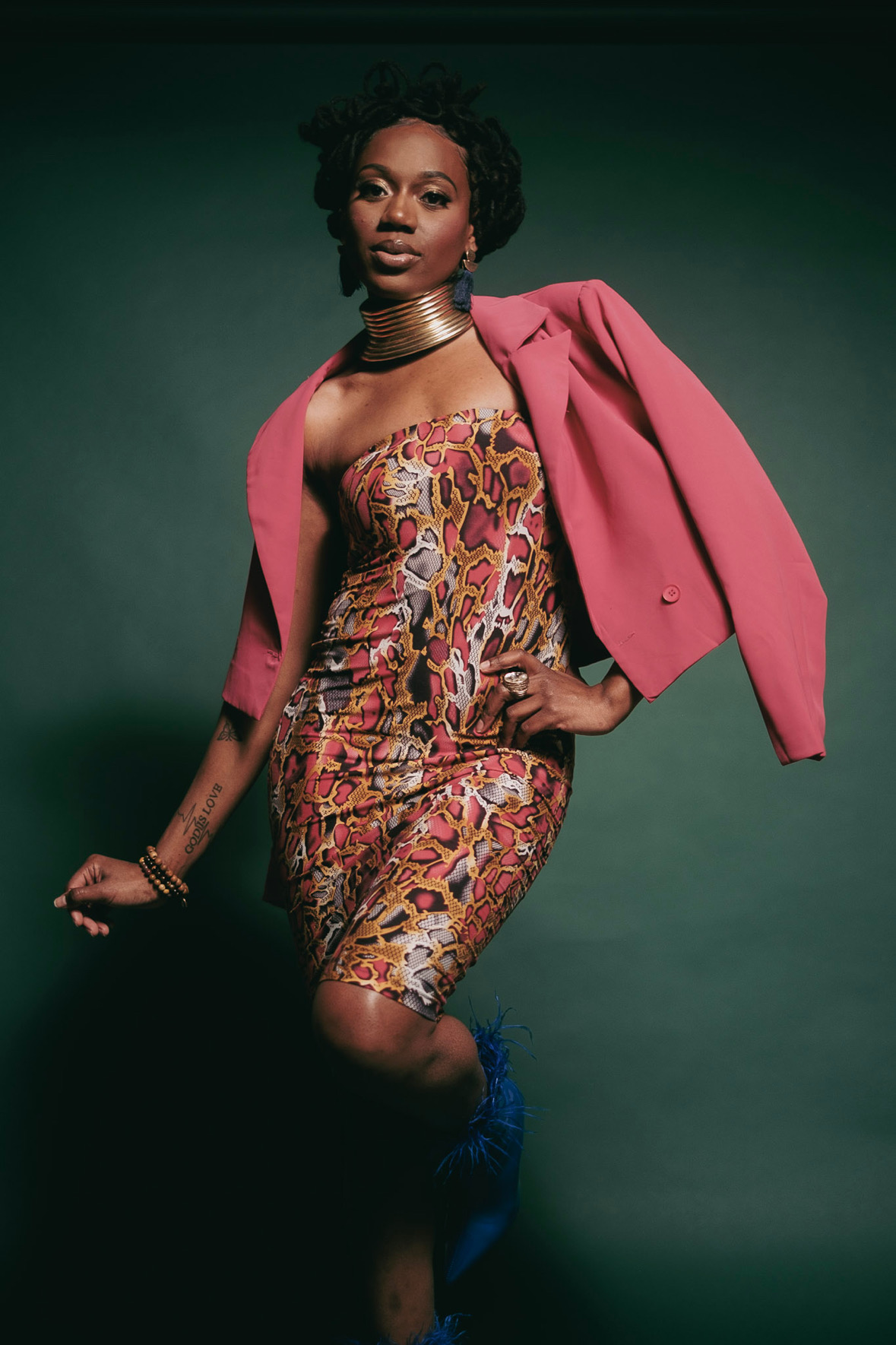 Woman in colorful dress with pink blazer draped over shoulders, standing against dark background.