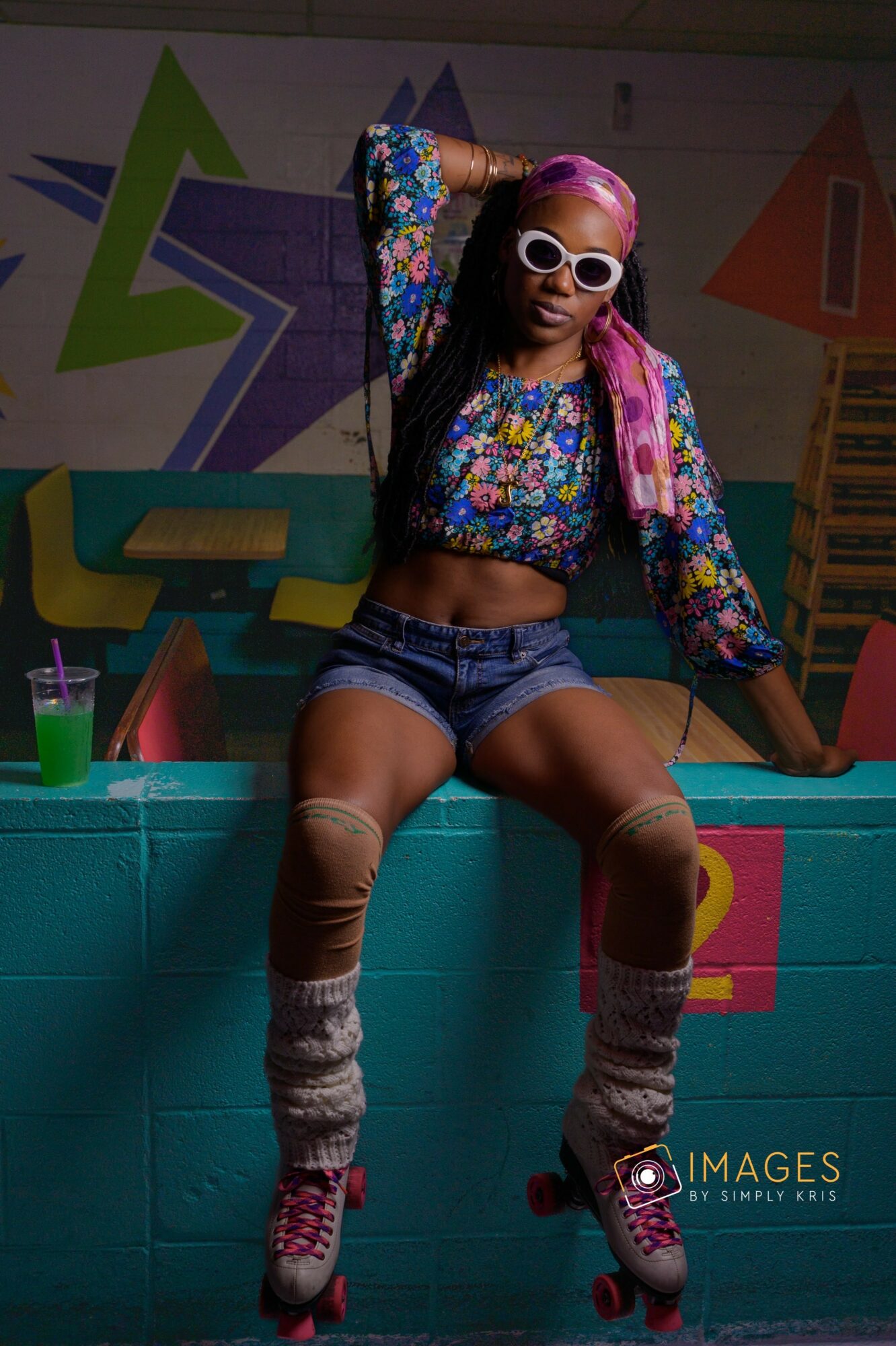 Person with pink hair, sunglasses, colorful jacket, crop top, shorts, and roller skates sitting on a ledge in front of a graffiti wall