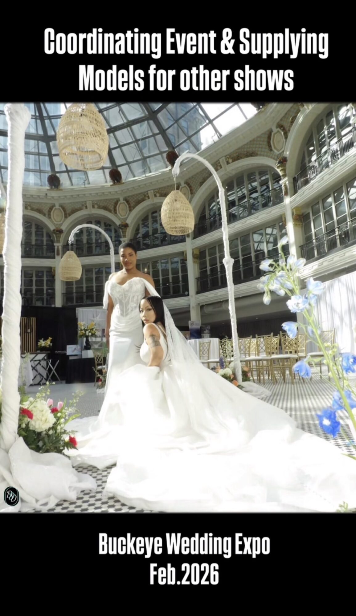 Bride and groom in a glass-ceiling venue with hanging lights and floral arrangements, bride in a long white gown, groom in suit.