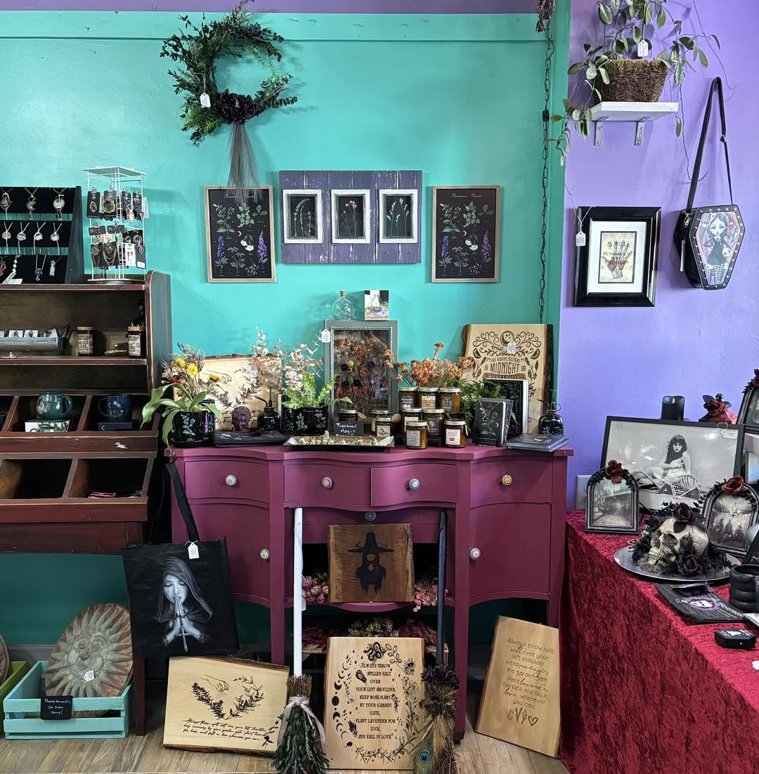 Display of framed artwork, jewelry, and decorative items on colorful tables and walls, with plants and accessories visible.