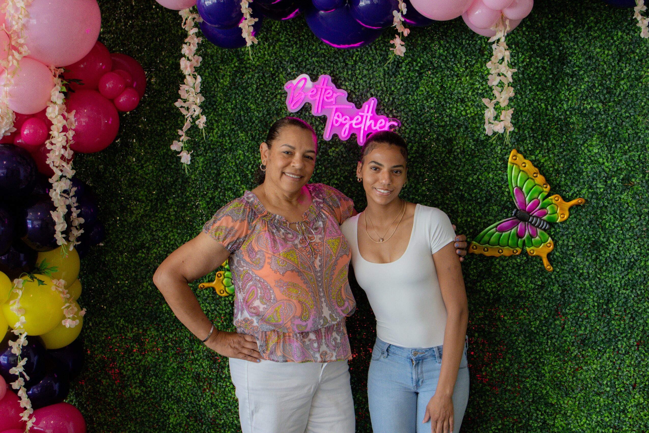 Two women stand together in front of a decorated backdrop with balloons, flowers, and a butterfly, smiling at the camera.