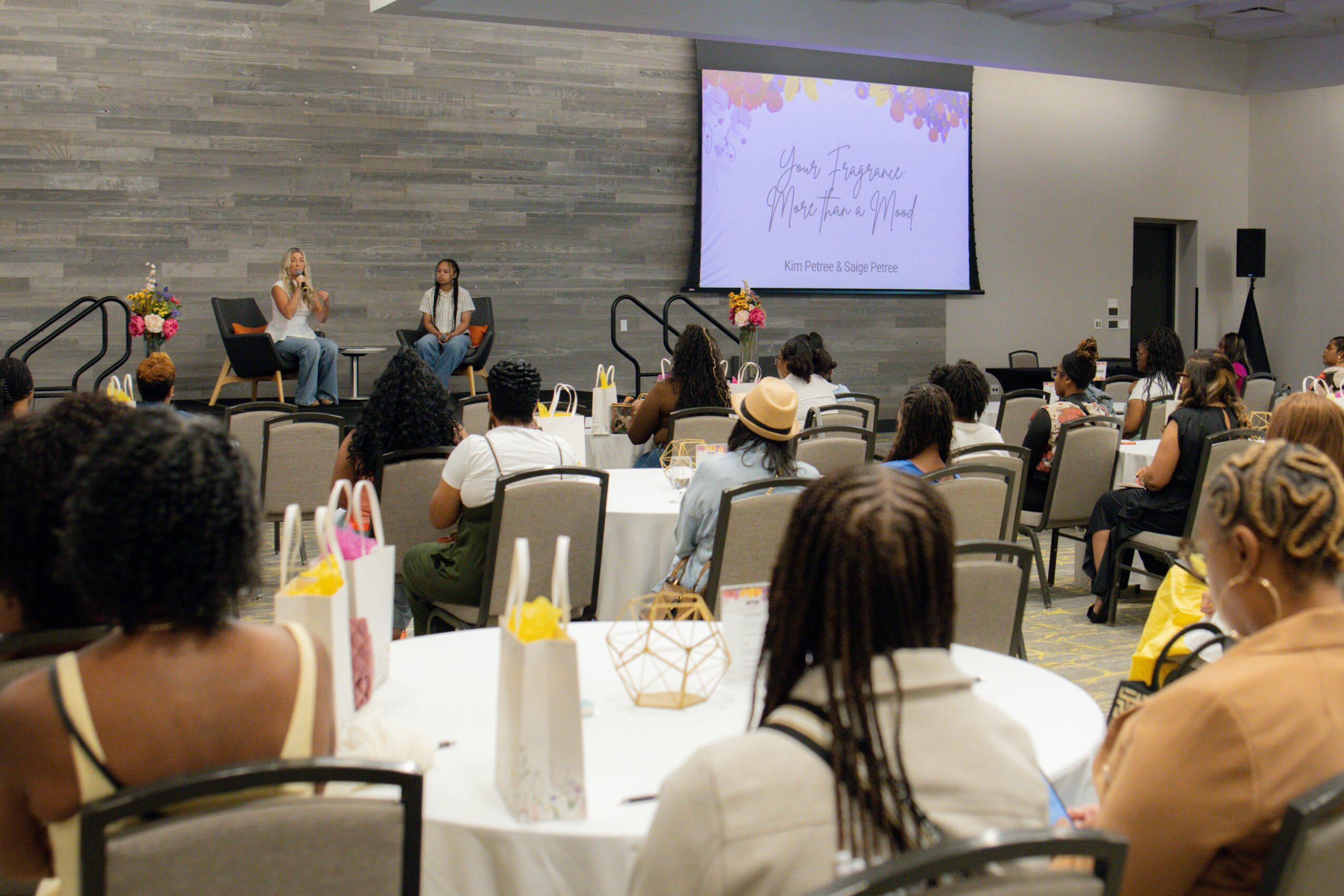 People seated at tables watching a panel of three women speaking on stage with a large screen behind them, in a conference room.