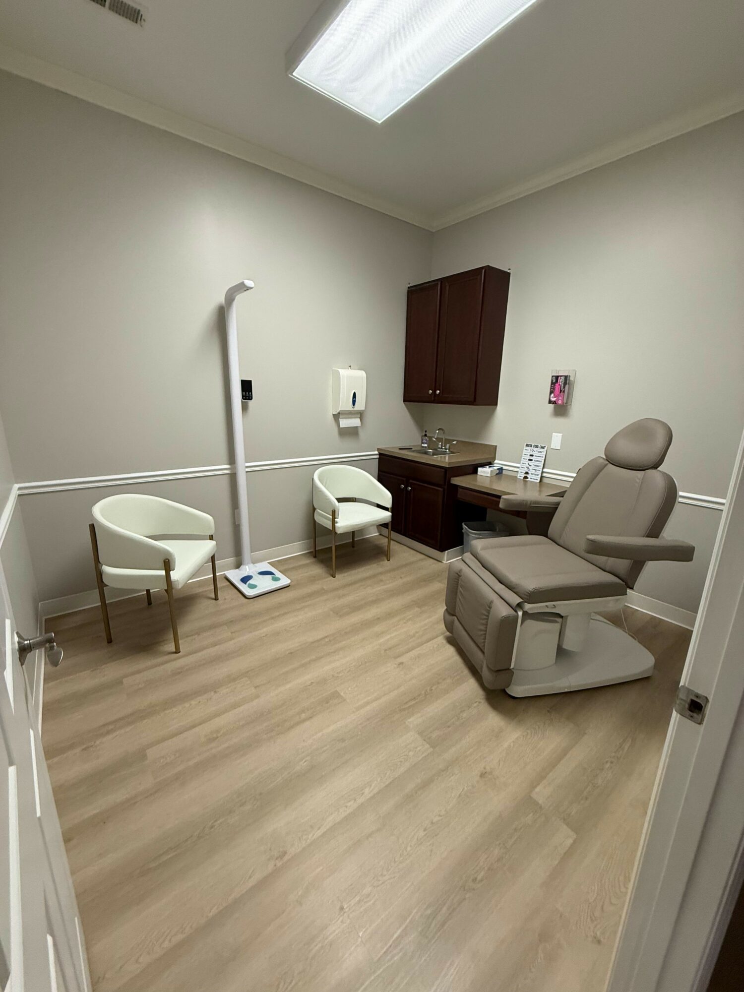 Upright hospital room with chair, cabinet, and medical equipment, light wood floor, white walls, and window with curtain.