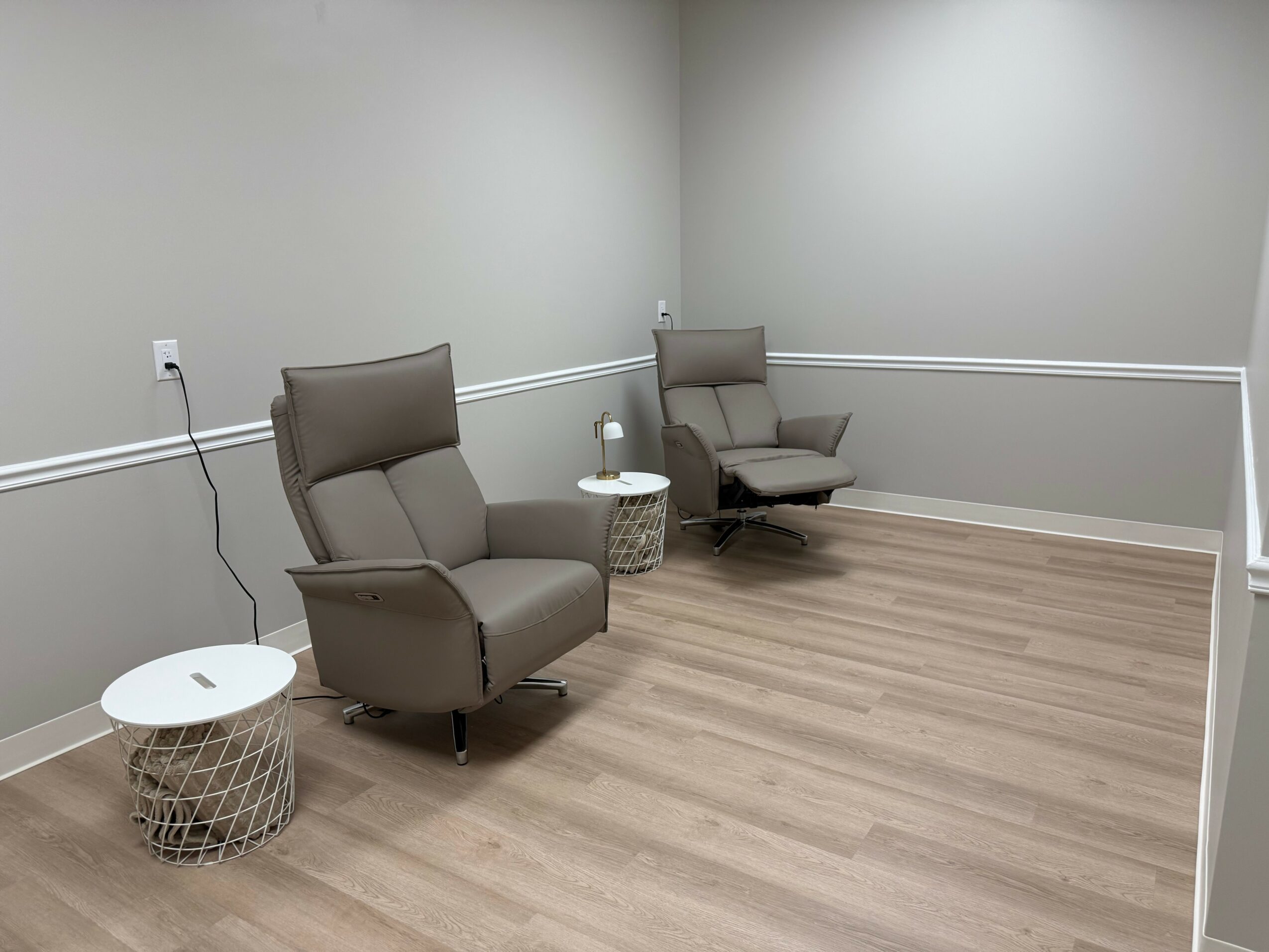 Two beige armchairs with small side tables in a plain room with light wood flooring and gray walls.