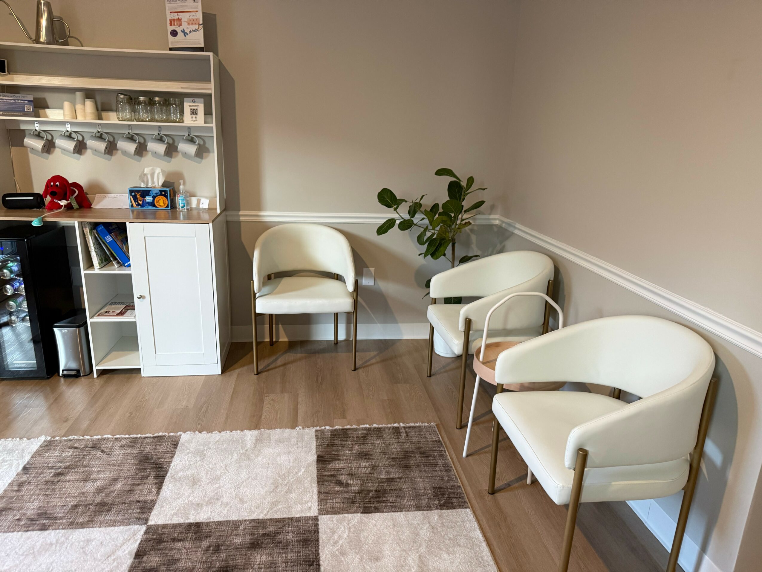 Waiting area with three white chairs, a potted plant, and a cabinet with glassware and toys, on wooden floor.