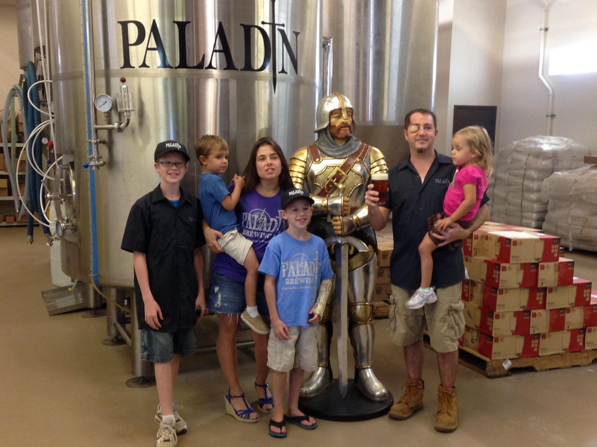 Group of children and adults with a knight statue inside a brewery, large brewing tanks in the background.