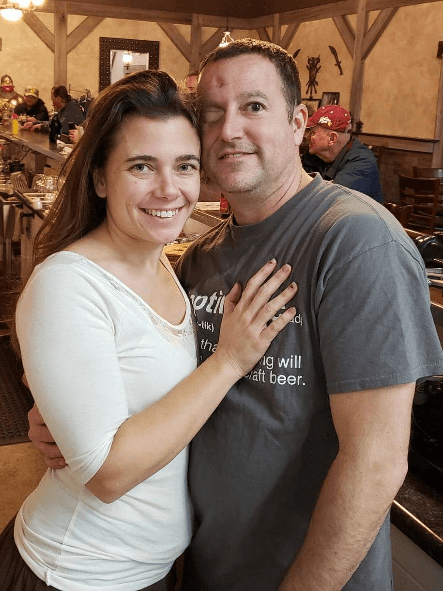A smiling woman with long brown hair and a man with short hair pose together indoors, woman’s hand on man’s chest.