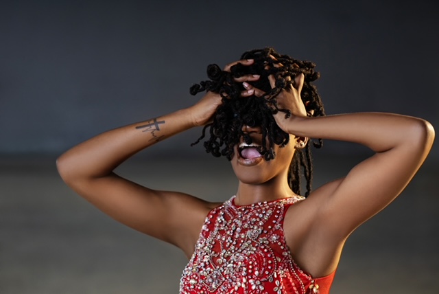 Person with curly hair covering face, wearing a red and silver dress, holding head with both hands.