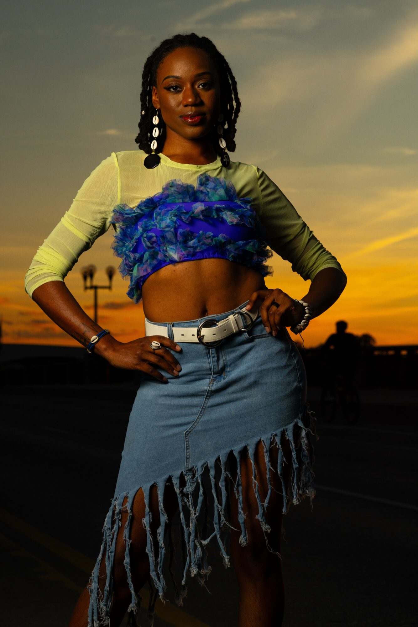 Woman with braided hair wearing a yellow top with blue feathered crop top and denim skirt at sunset
