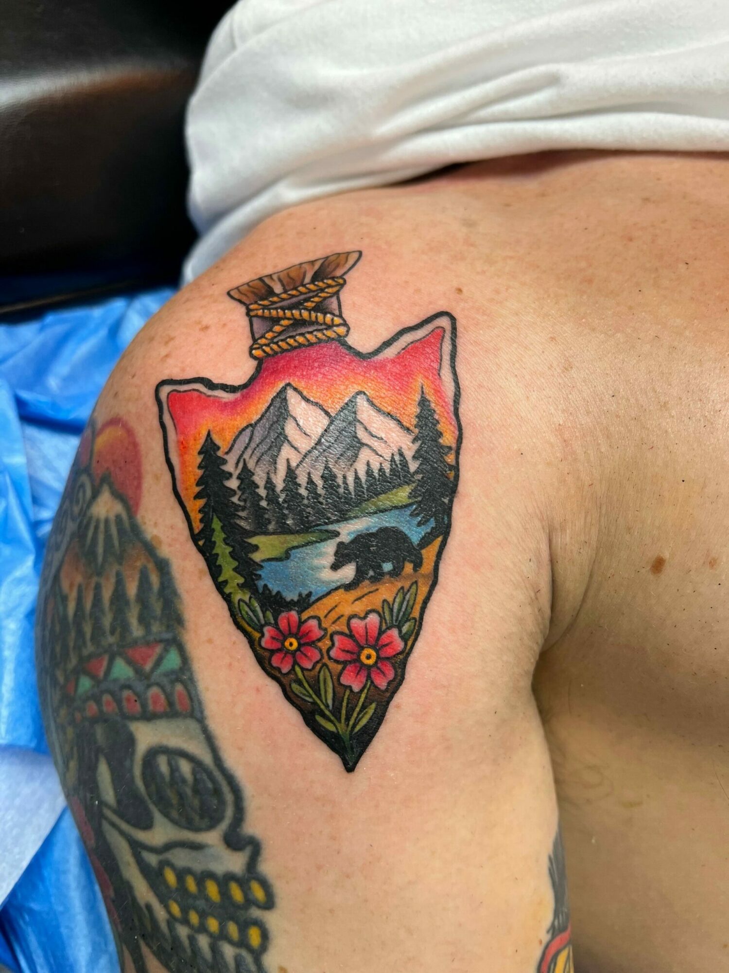 Colorful tattoo of mountains, trees, and flowers on person's upper arm, with a butterfly nearby, against a light skin background.