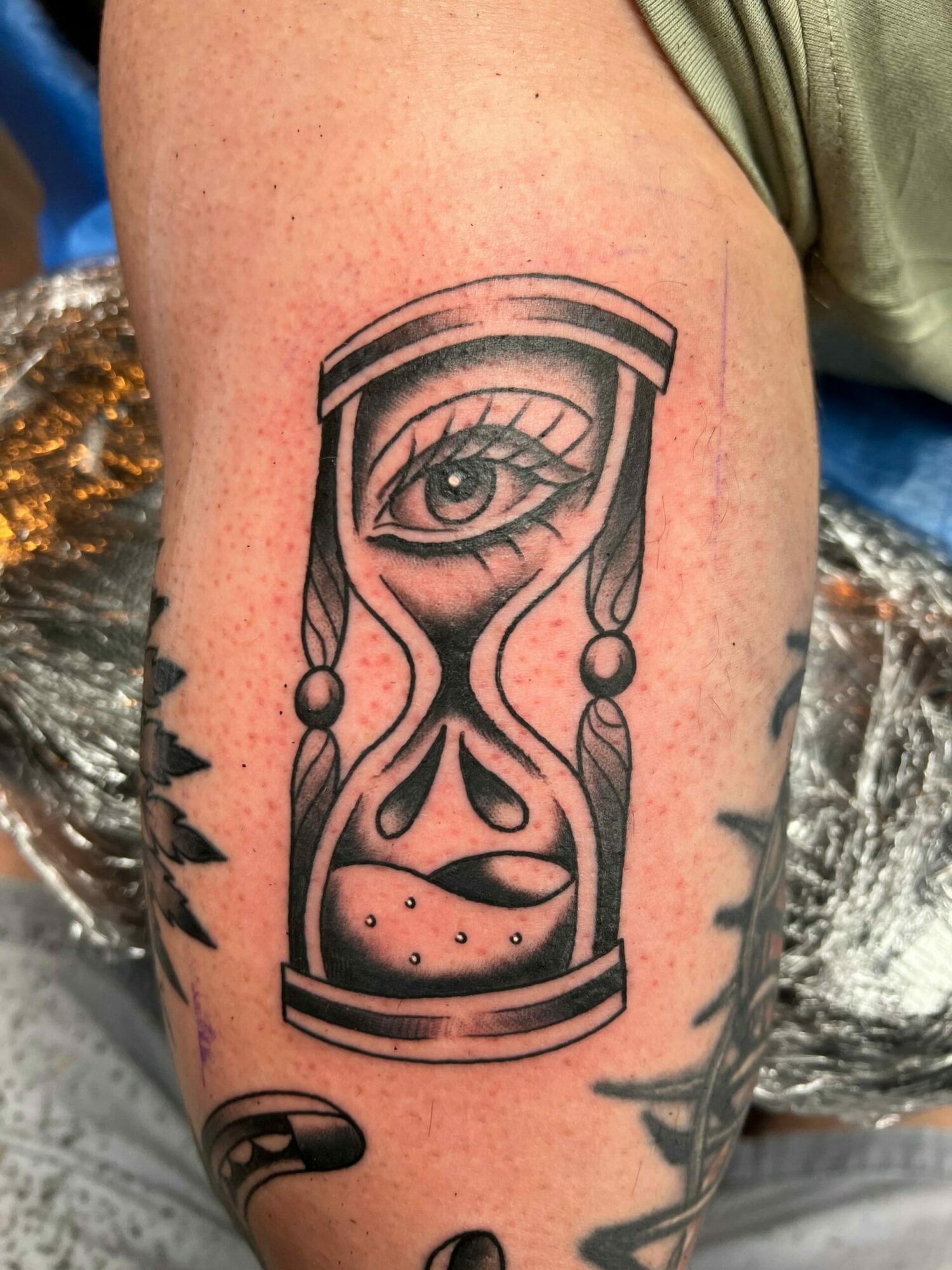 Tattoo of a stylized hourglass with an eye inside, on a person's arm, with a sun design above.