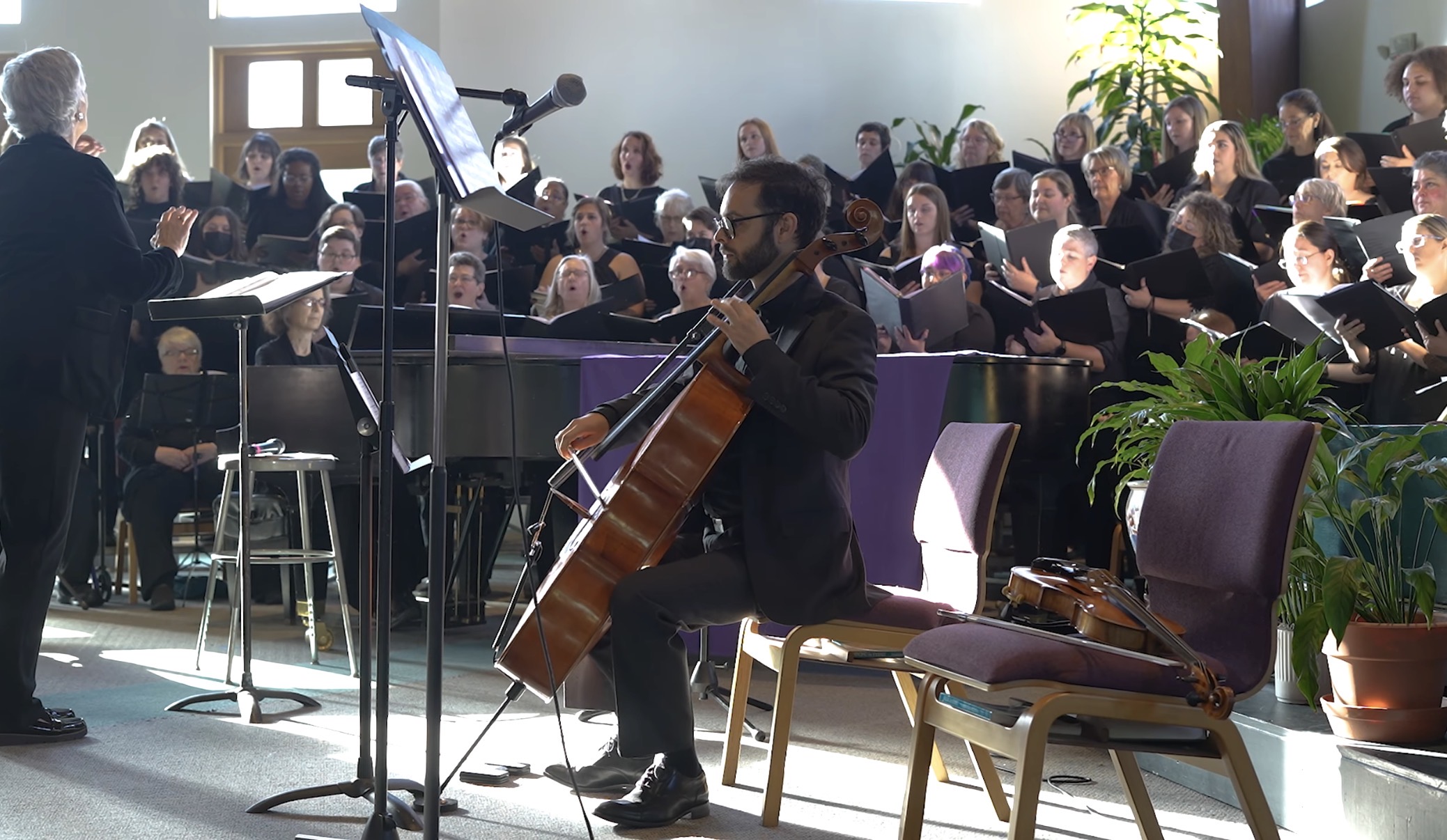 A man playing guitar in front of a choir during a performance, with a conductor nearby and audience in background.