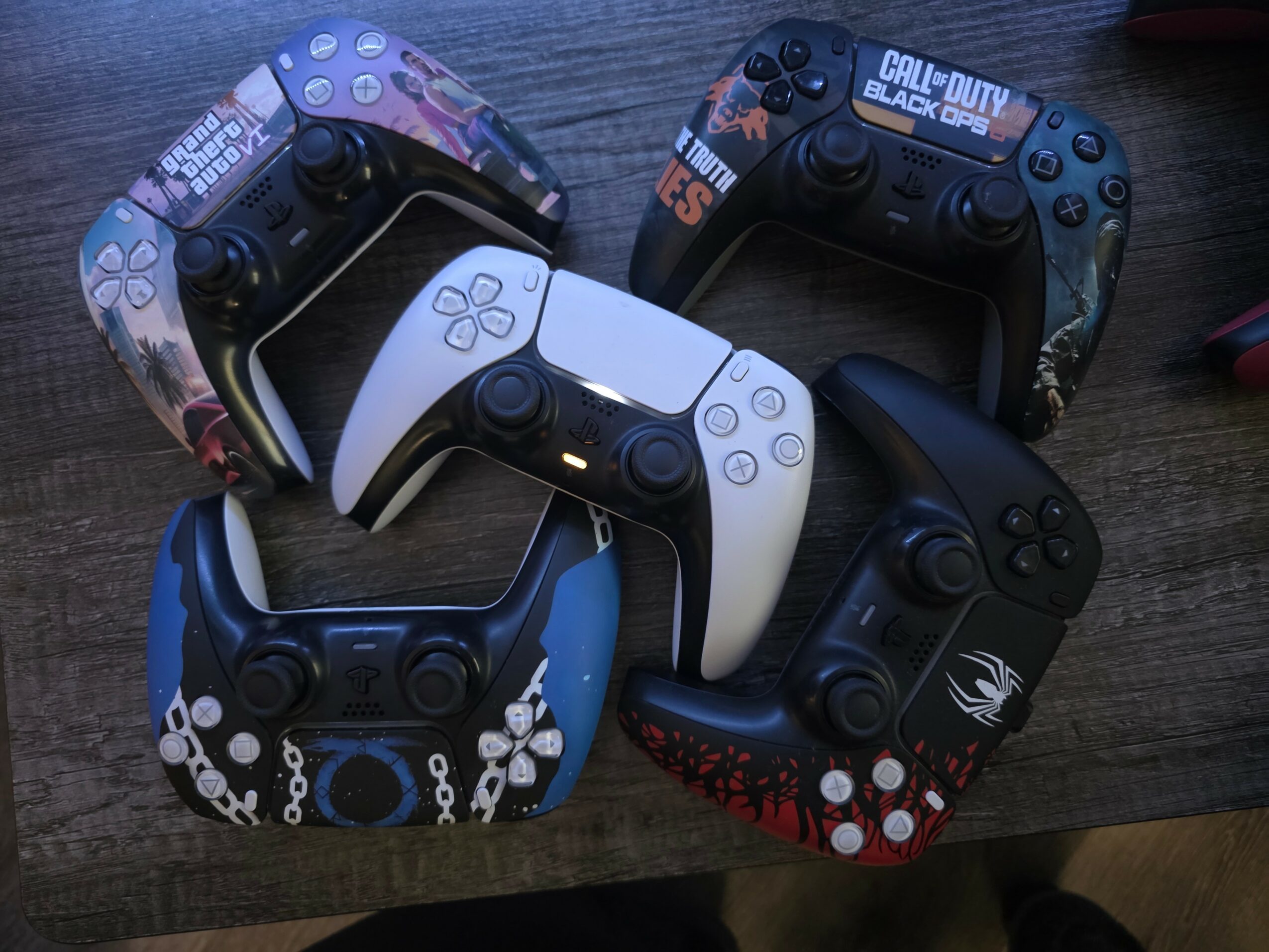 Six gaming controllers arranged on a wooden surface, with various colors and designs, including black, white, blue, and patterned styles.
