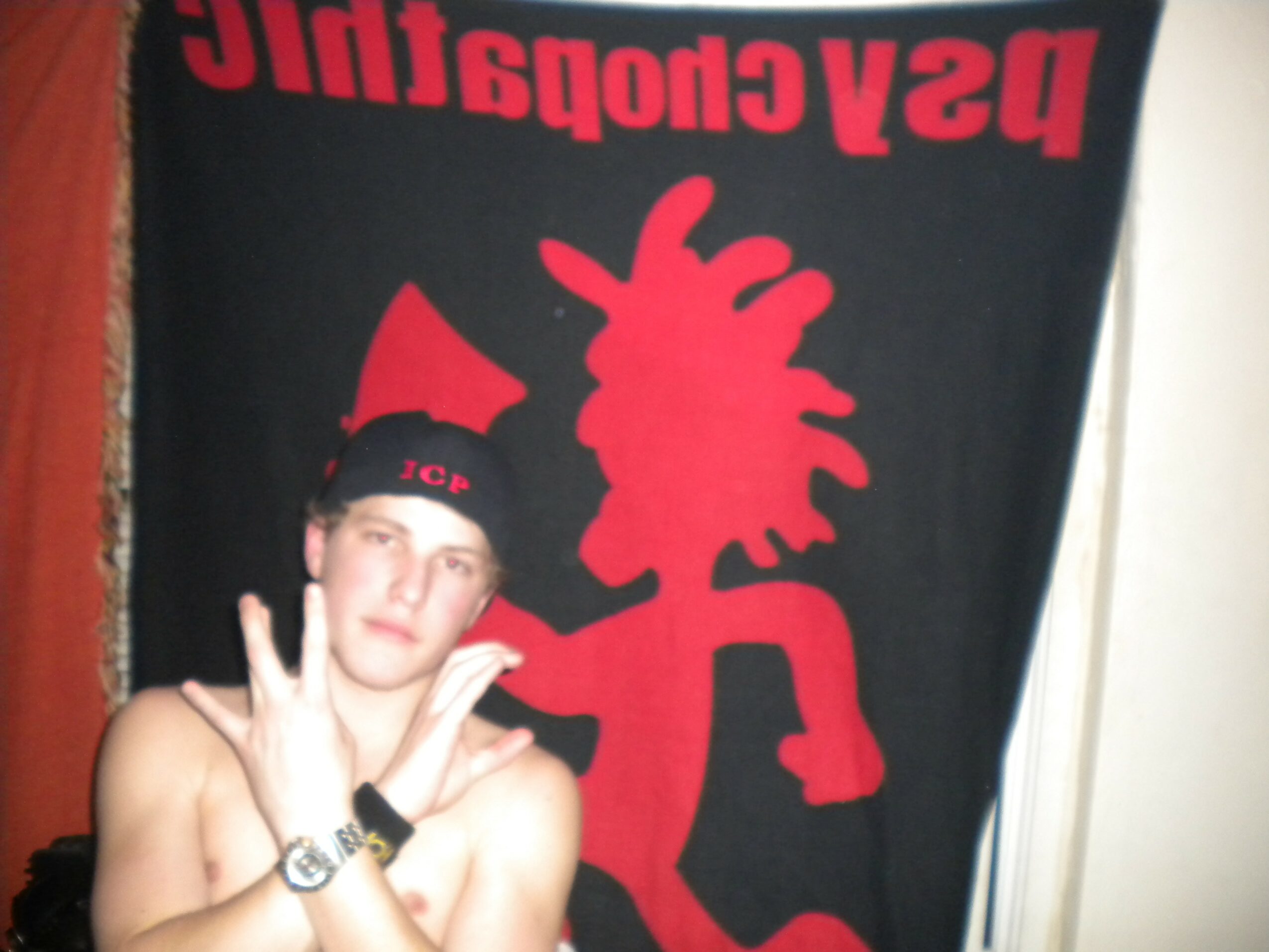 Young person with short hair making peace signs, wearing a black hat, in front of a red and black banner.