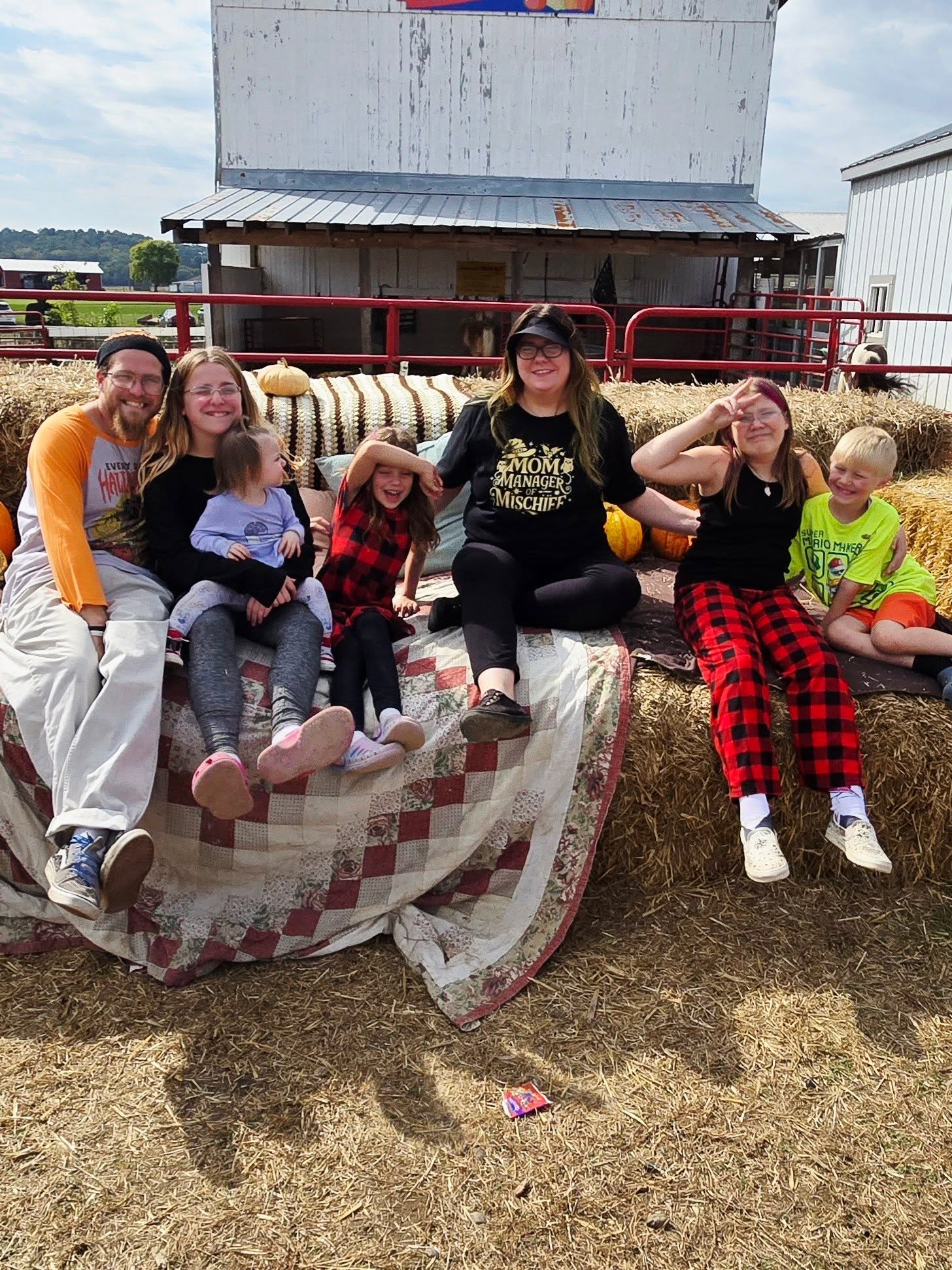 Group of seven people sitting on a hay bale in front of a farm building, smiling outdoors on a sunny day.