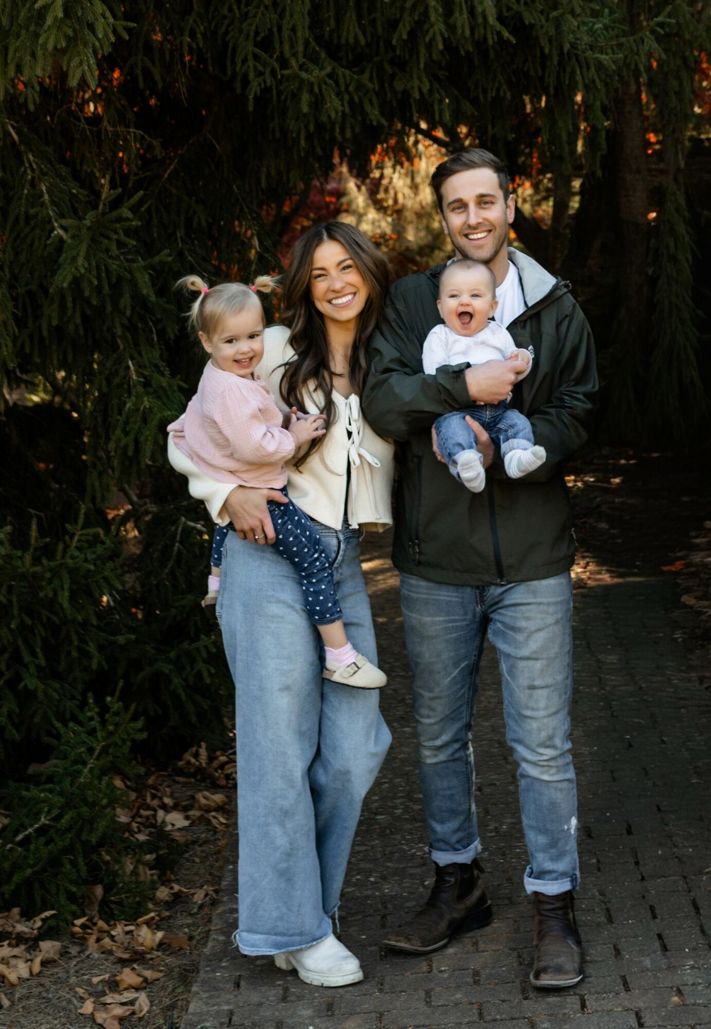 Family of four outdoors, two adults and two children, smiling, standing on a path with trees in background.