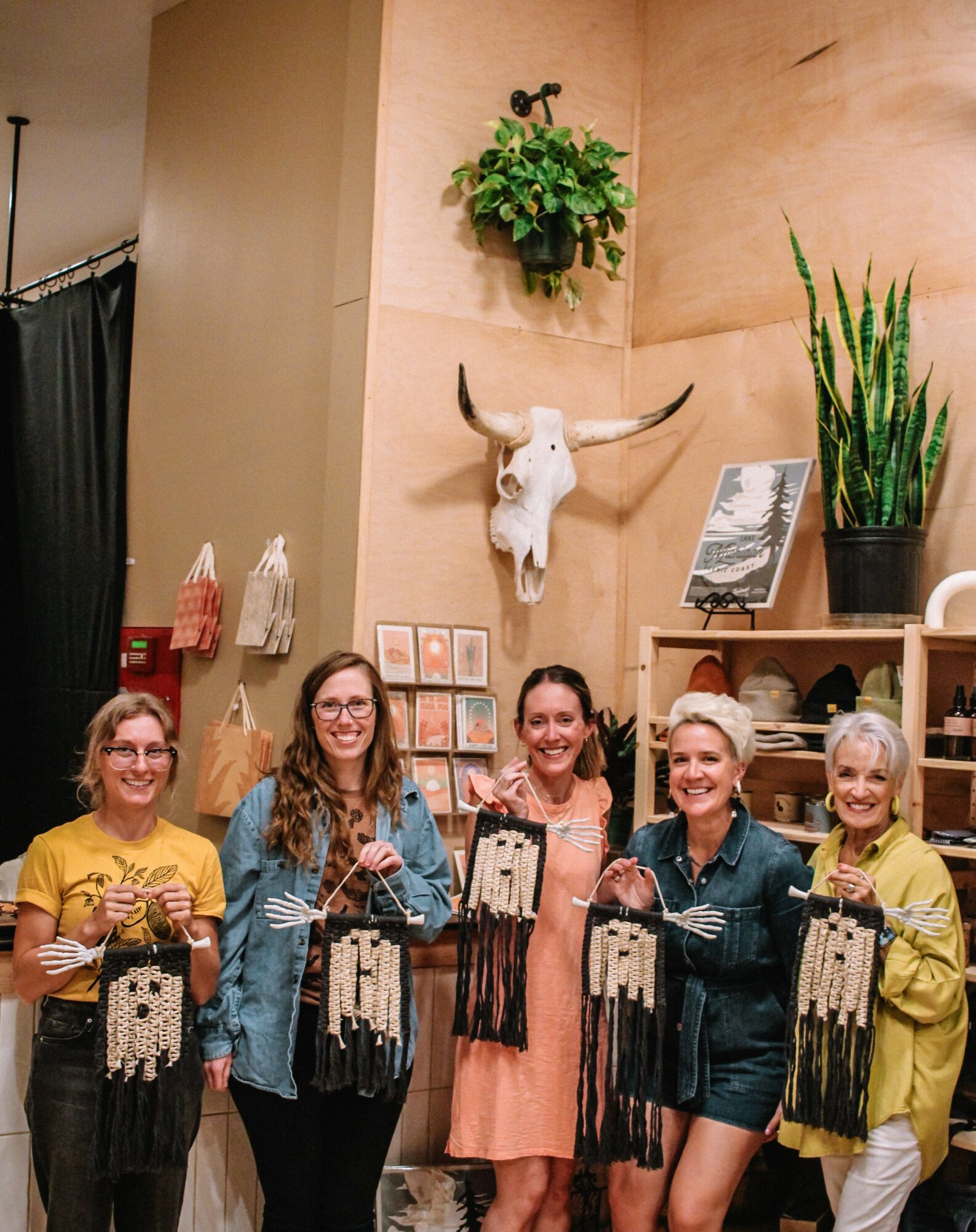 Five women stand in front of a wall with a mounted animal skull, potted plants, and framed pictures, smiling and holding handmade crafts.