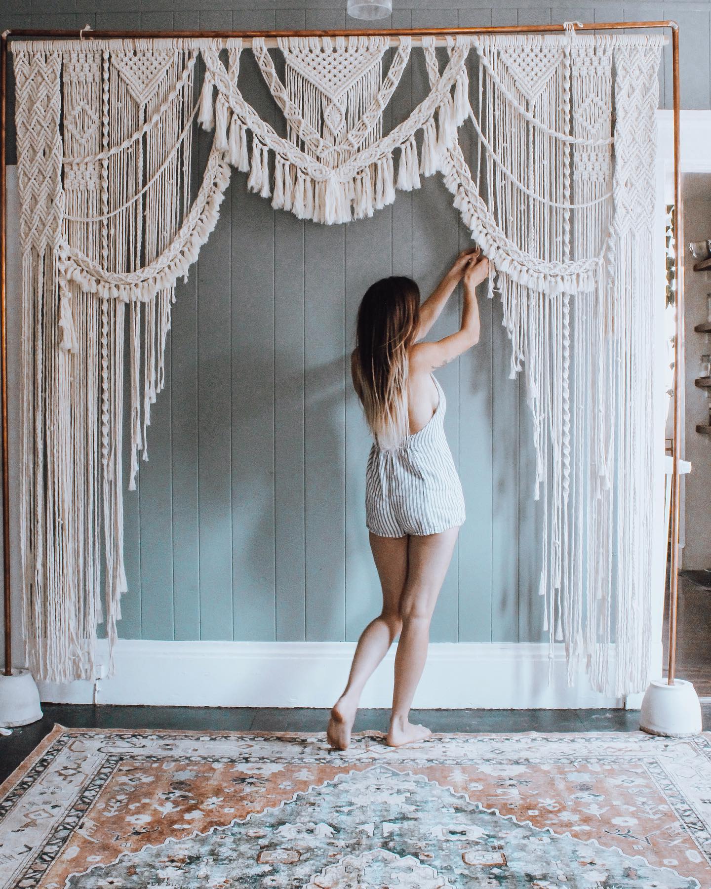Woman with long hair in a white outfit adjusting decorative hanging curtains on a wall, standing on a patterned rug.