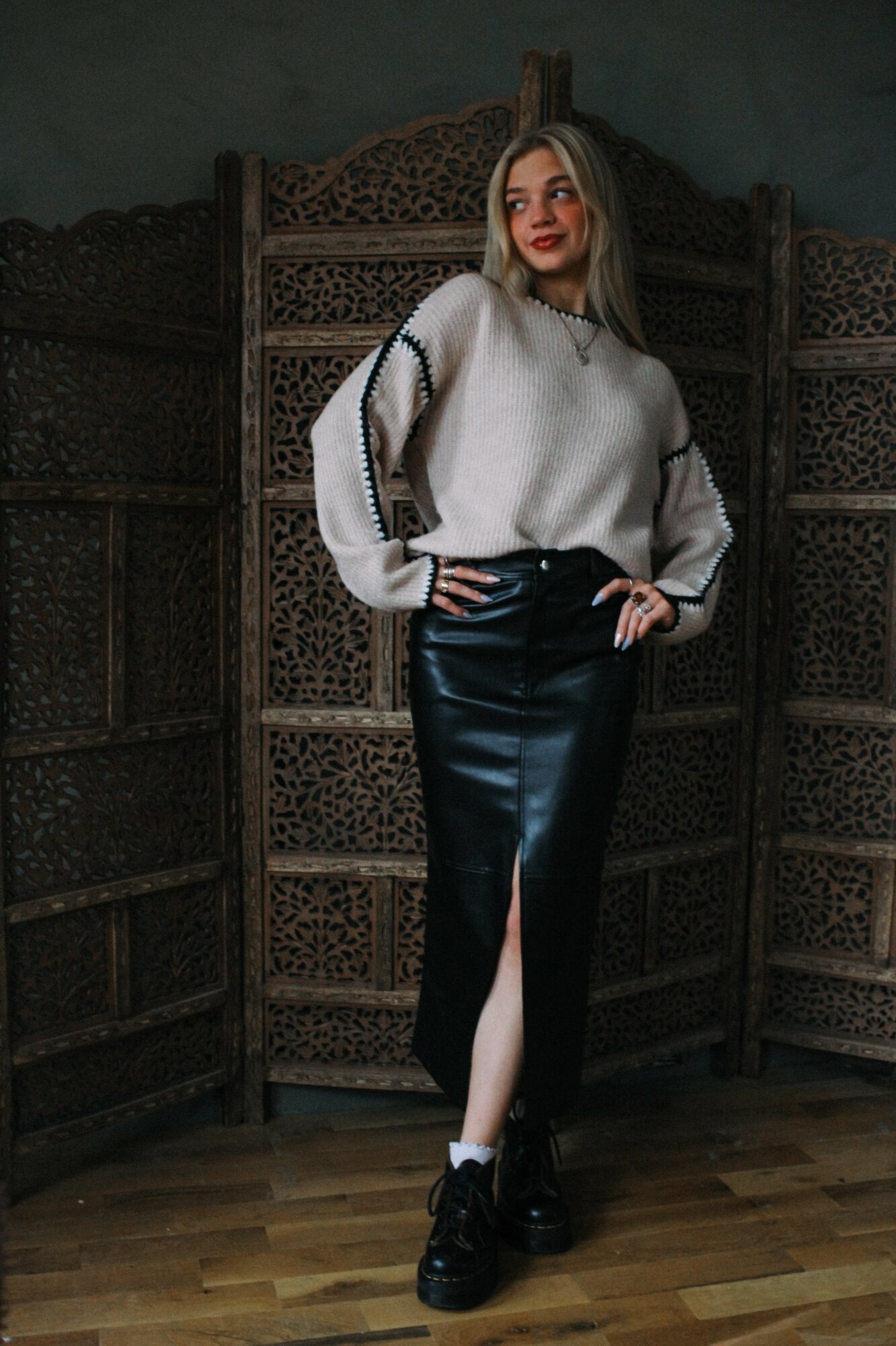 Woman with long hair wearing a white sweater and black skirt with a slit, standing indoors in front of a wooden screen.