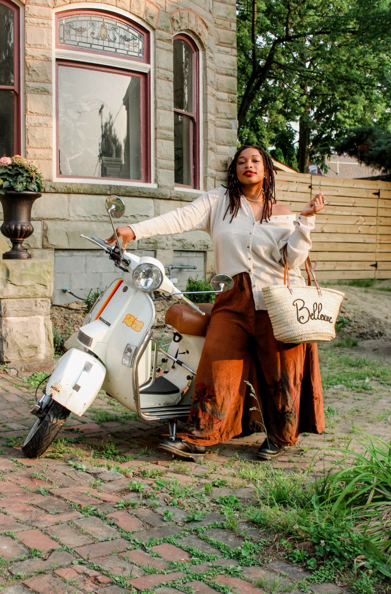 Woman standing next to a white scooter outdoors, holding a bag, with a building and trees in the background.