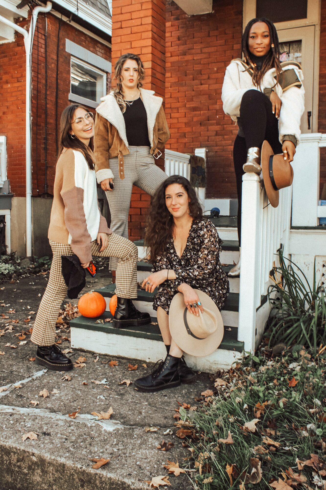 Four women pose on a porch with pumpkins, dressed in fall clothing, in front of a brick house.