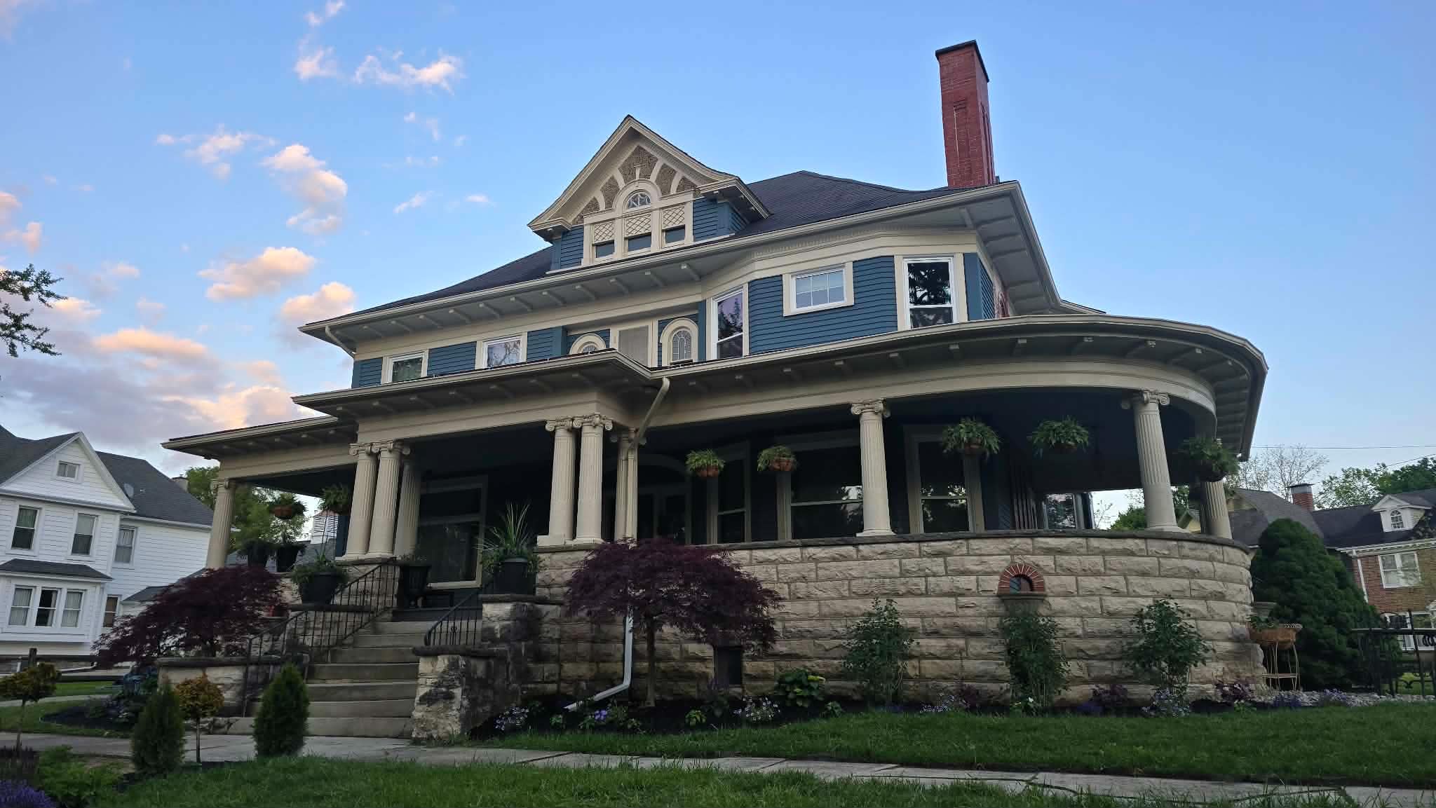 Large Victorian-style house with a wraparound porch, multiple gables, and a chimney, set against a blue sky with clouds.