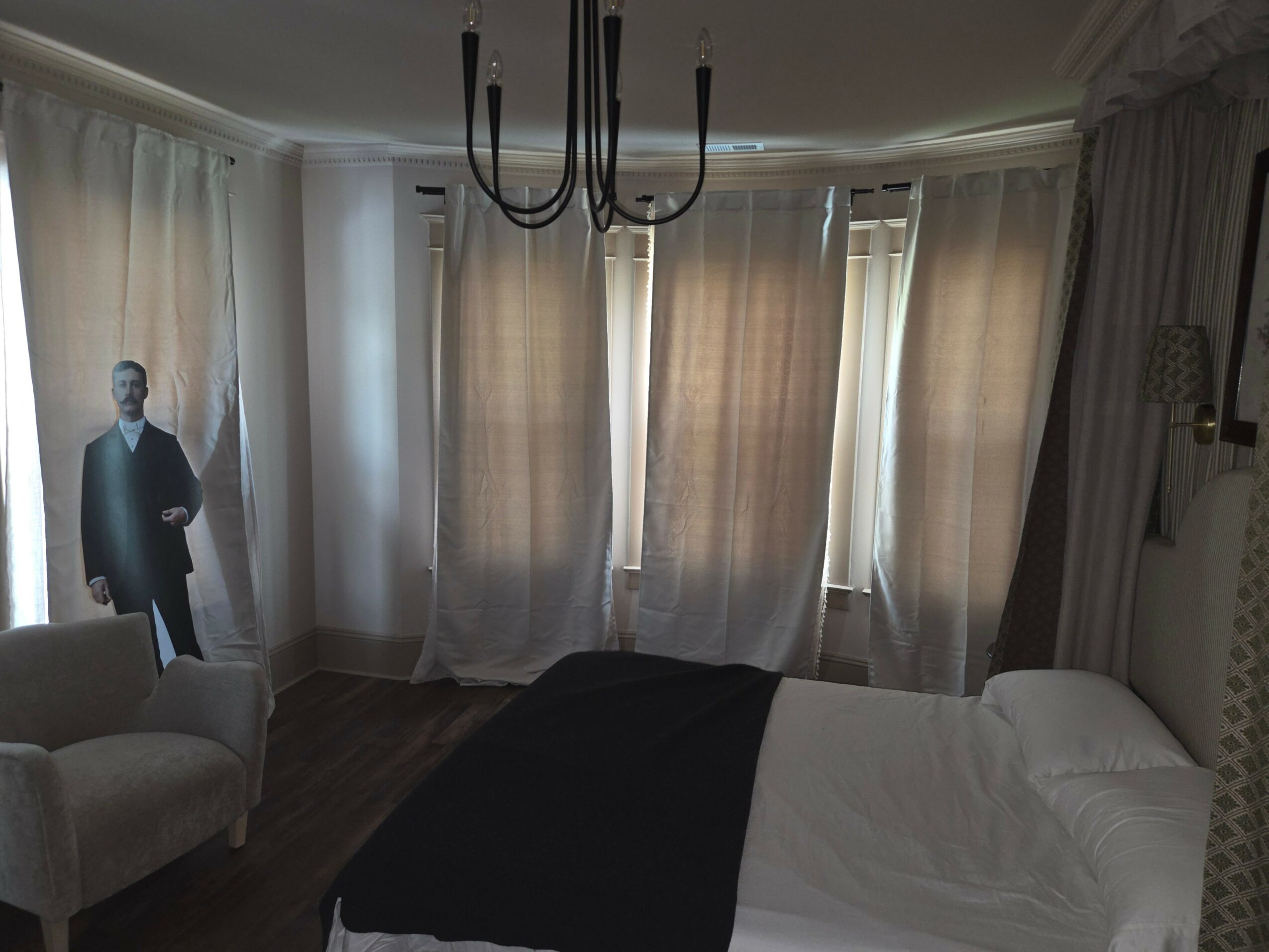 Room with a bed, curtains, chandelier, and a life-sized cutout of a man in a suit, near a chair.