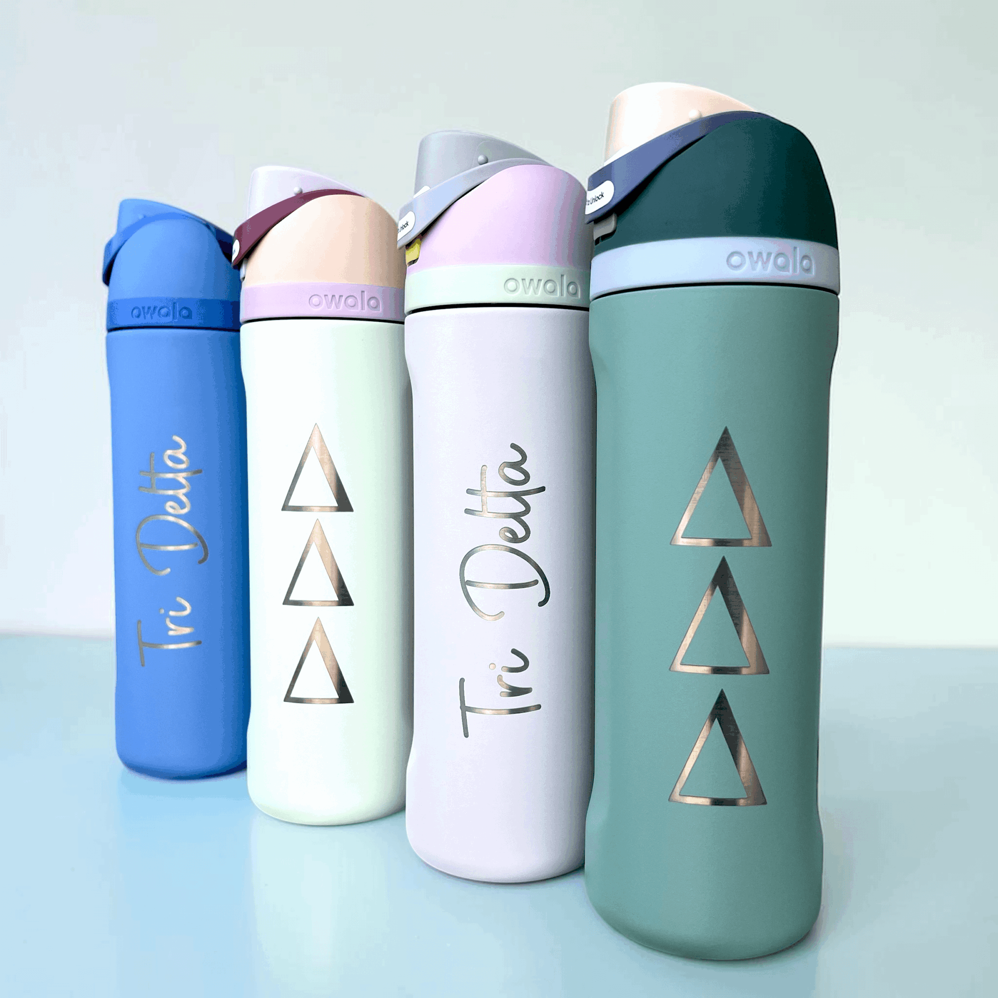 Four reusable water bottles in blue, white, light purple, and green, with geometric triangle designs, on a light background.