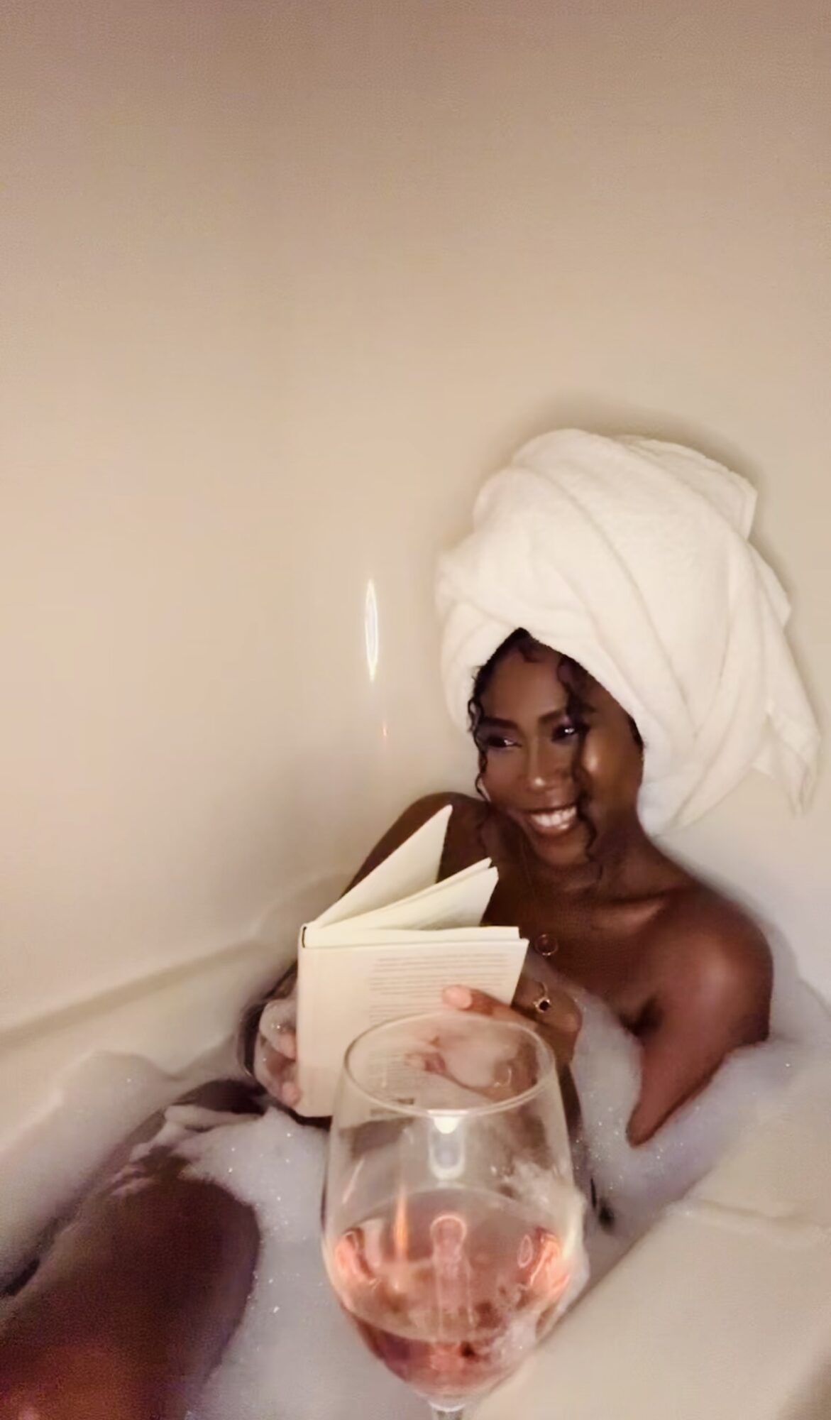 Person with towel on head reading a book in bath, holding a glass of pink drink, smiling.