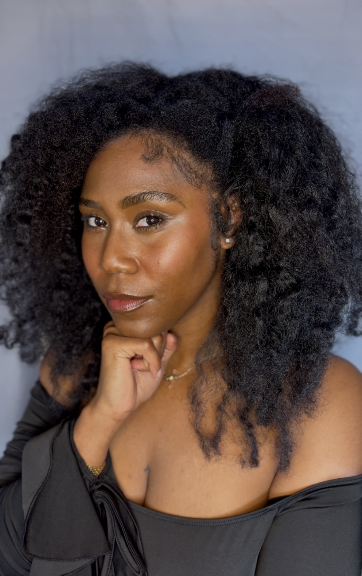 Portrait of a woman with voluminous curly hair, resting her chin on her hand, wearing a black off-shoulder top.