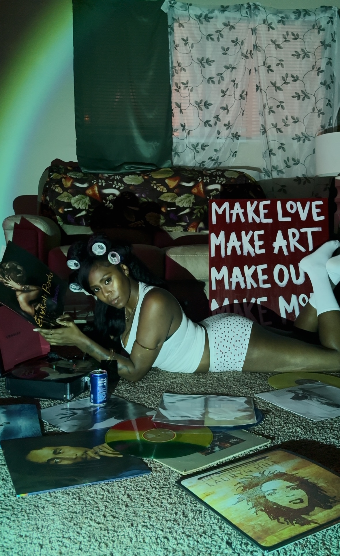 Person lying on the floor with a laptop, surrounded by books and papers, in a room with curtains and a sign.
