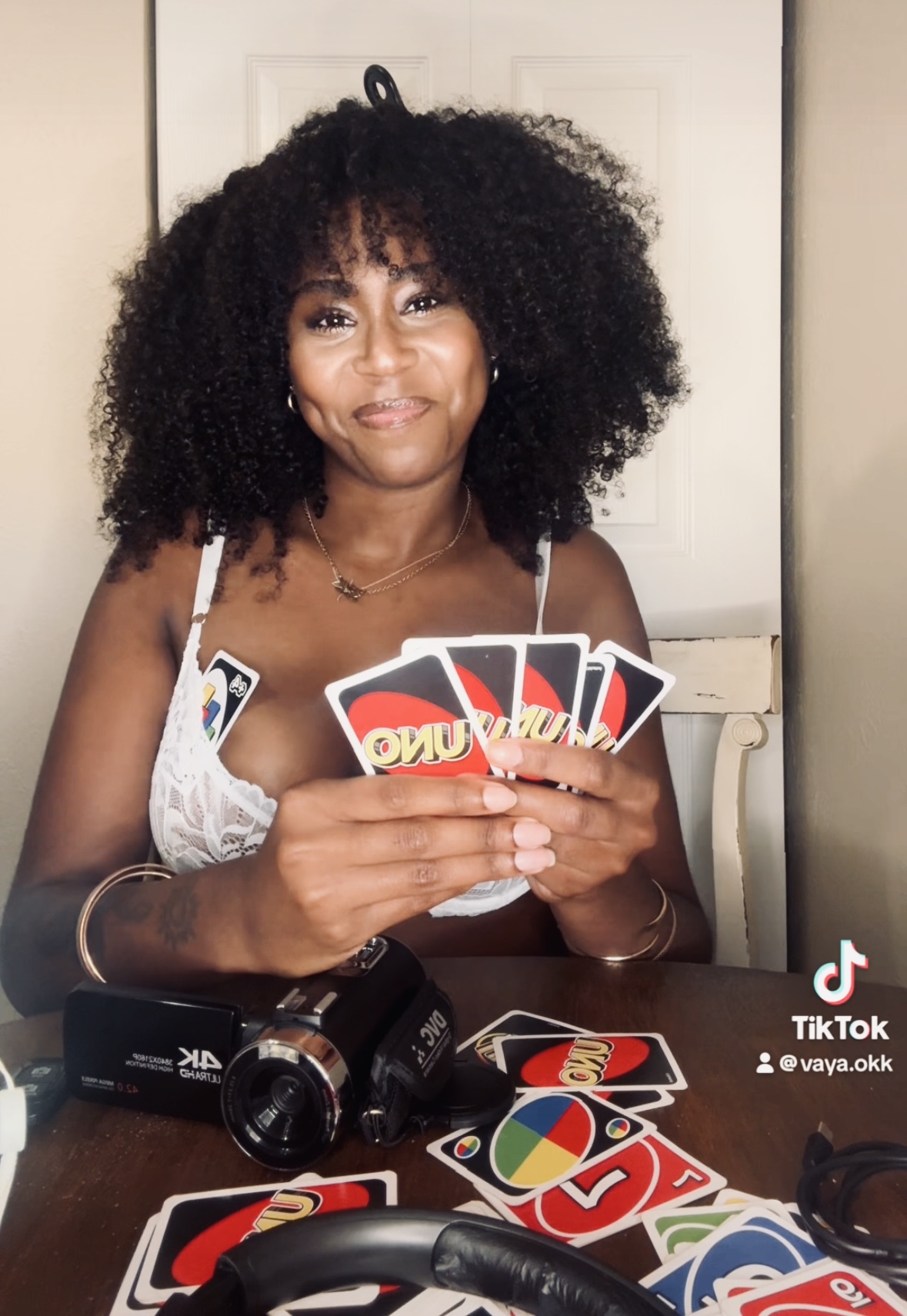 Woman with curly hair holding playing cards, sitting at a table with a camera and more cards, smiling.