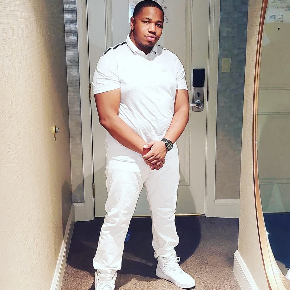 Young man in white outfit standing indoors with arms crossed, in front of a door and mirror.