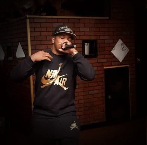 Person wearing a Nike Air sweatshirt and cap, holding a microphone, standing in front of a brick wall.