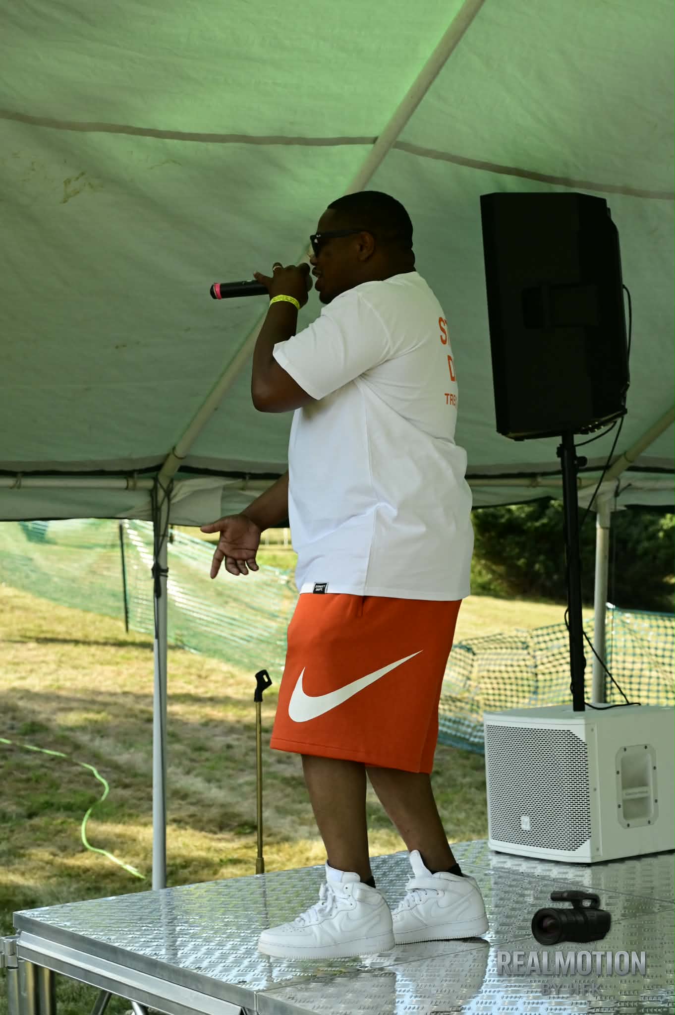 Person wearing white shirt and orange Nike shorts holding a microphone, standing on a platform under a tent outdoors.