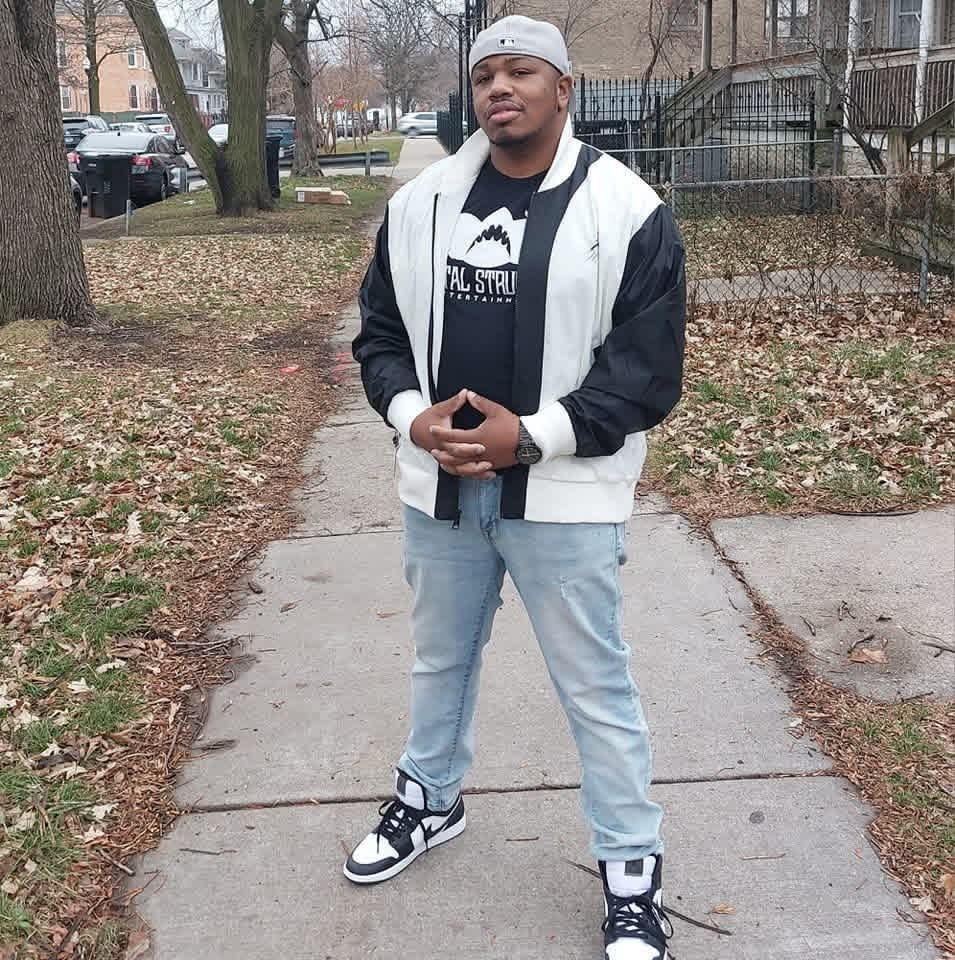 Young man standing on a sidewalk outdoors, wearing a white and black jacket, light blue jeans, and sneakers, with trees and buildings in background.