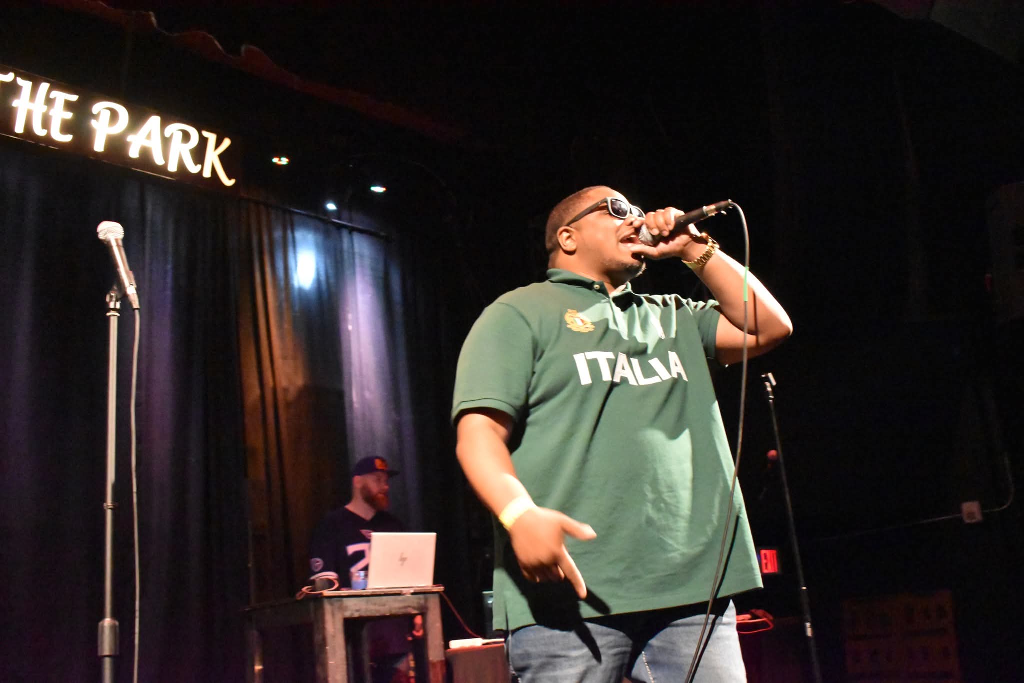 Performer singing into microphone on stage with a person working at a laptop in the background.