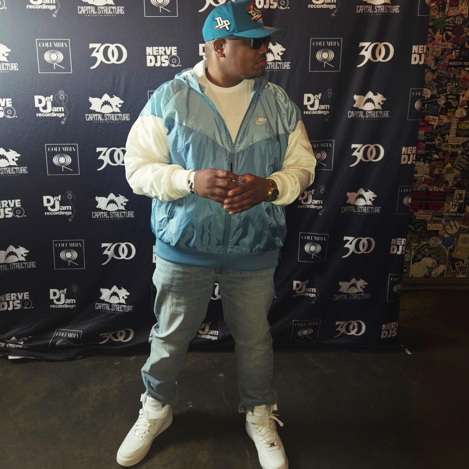 Person wearing a blue cap, sunglasses, light-colored jacket, and jeans standing in front of a black backdrop with logos.