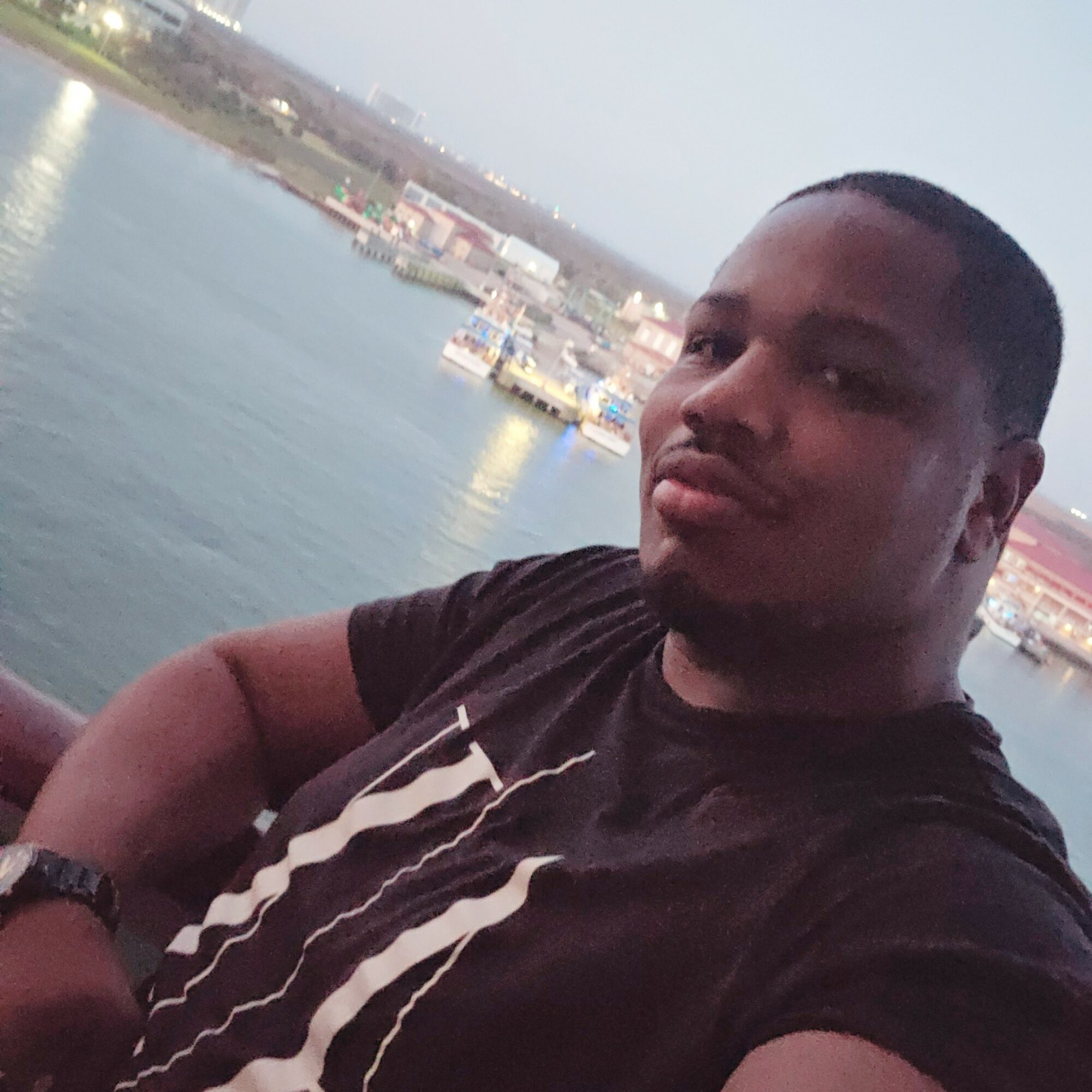 Young man with short hair and a black T-shirt taking a selfie near water with boats and buildings in the background.