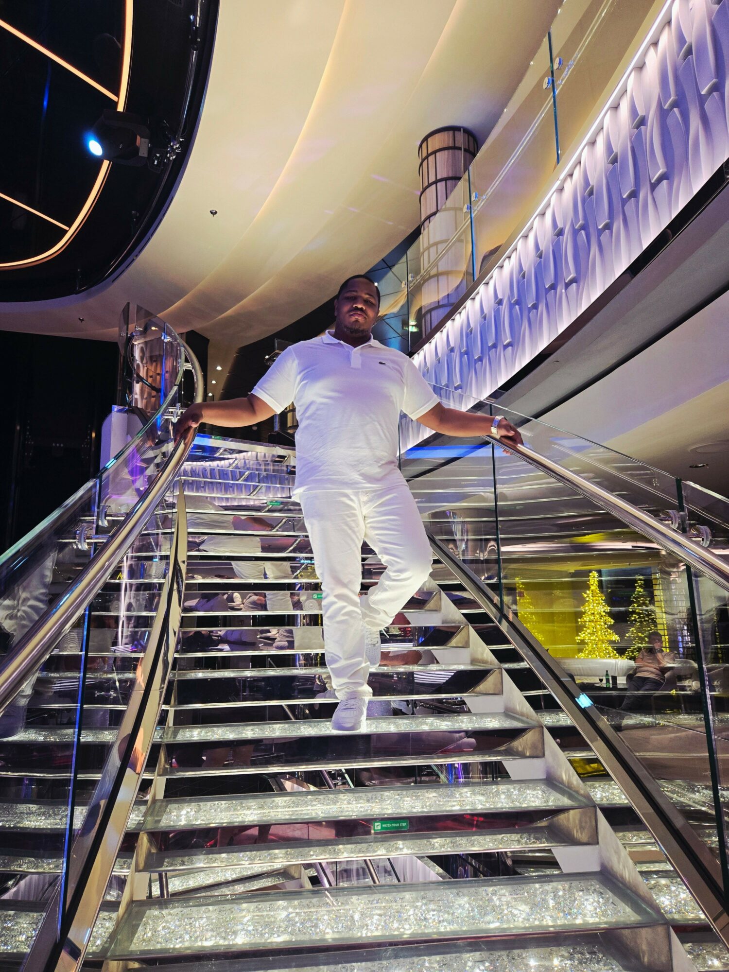 Person in white outfit leaning on a railing inside a modern building with colorful lighting, glass walls, and reflections.