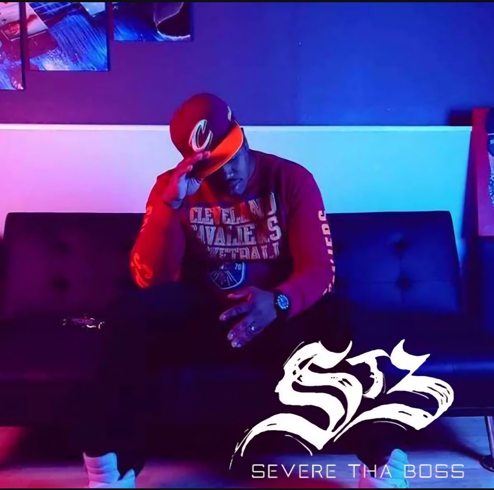Person sitting on a black couch in a dimly lit room with blue and purple lighting, wearing a red cap and jacket, holding a drink.
