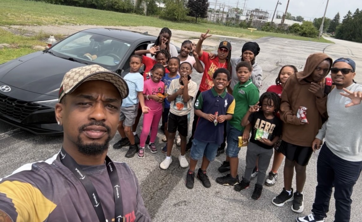 Group of children and adults posing outdoors near a black sports car, with a man taking a selfie in the foreground.