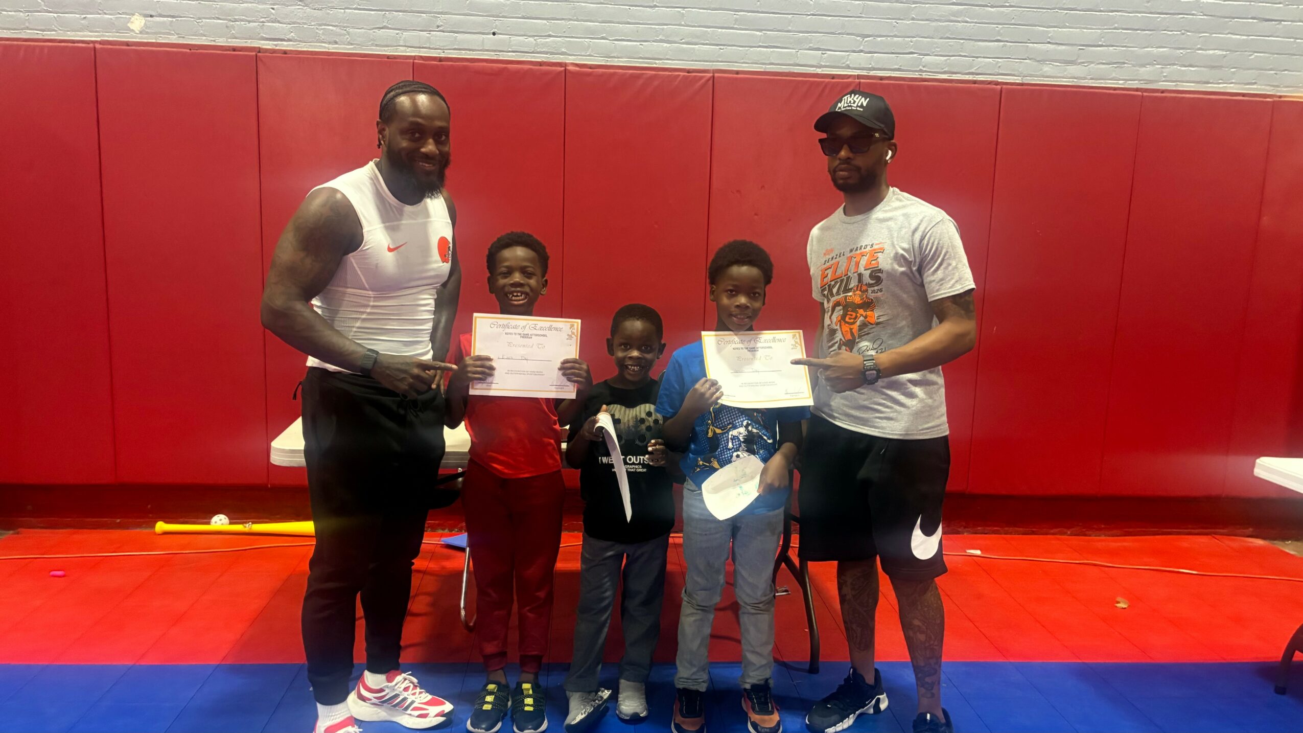 Two adults and three children standing on a gym floor holding certificates, with a red padded wall behind them.