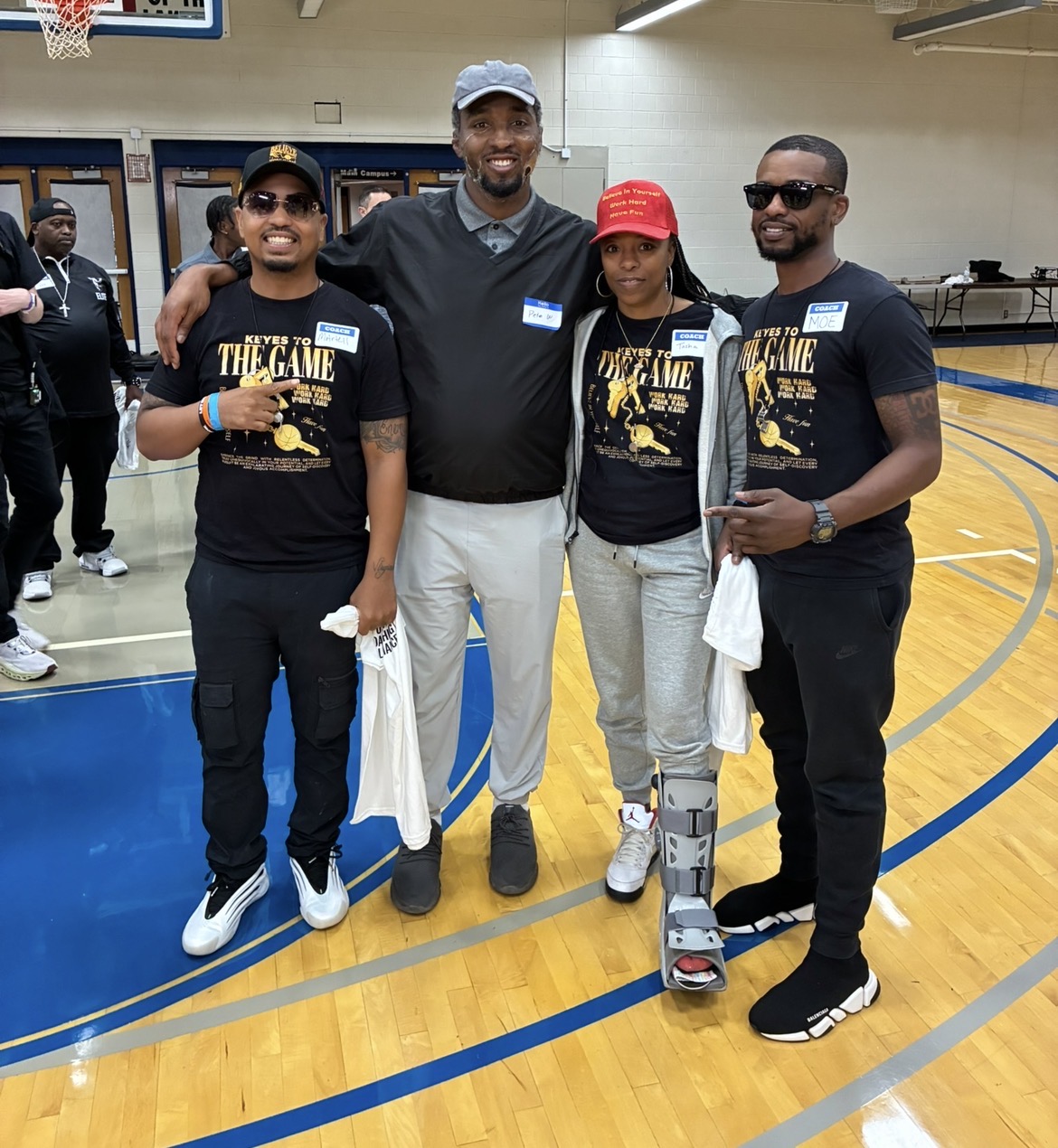 Four people standing on a basketball court, smiling, with two wearing matching black T-shirts, one in a red cap, and one with a medical brace.