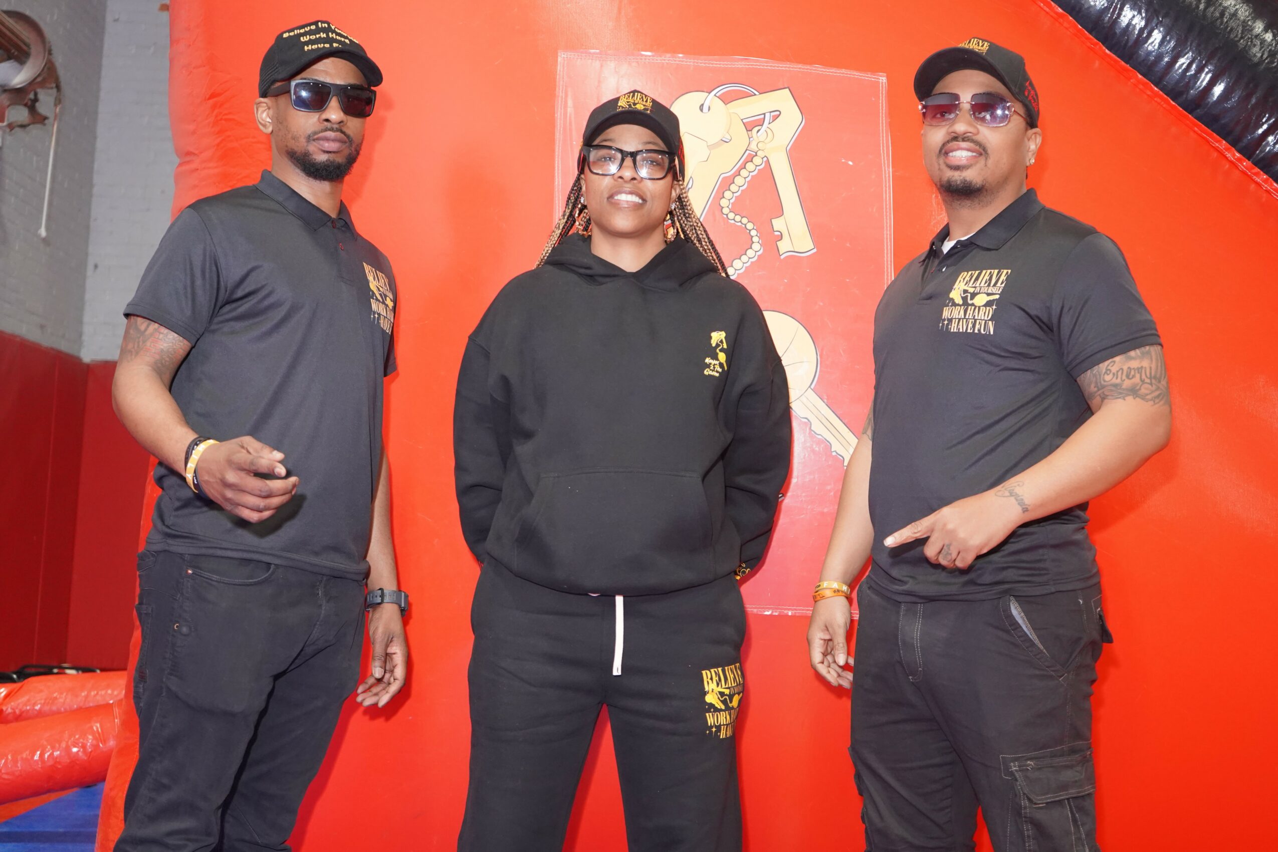 Three people standing together in front of a red background, smiling, wearing black clothing and sunglasses.