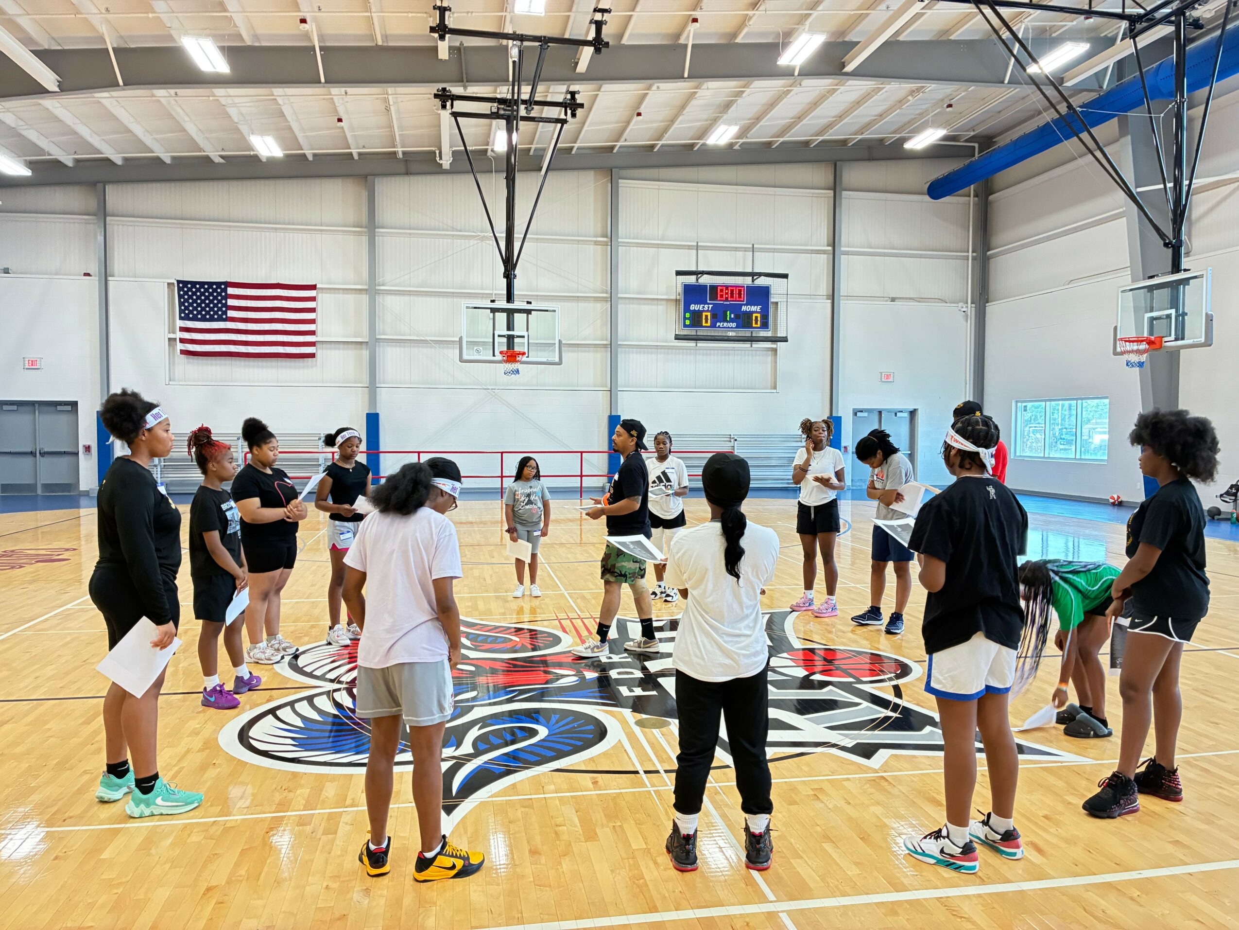 Group of children and adults standing in a circle on a basketball court inside a gymnasium, with an American flag on the wall.